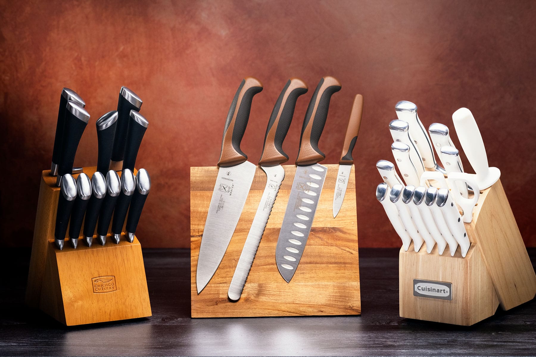 Three of the best budget kitchen knife sets under $100 sitting on a countertop in front of a maroon background.