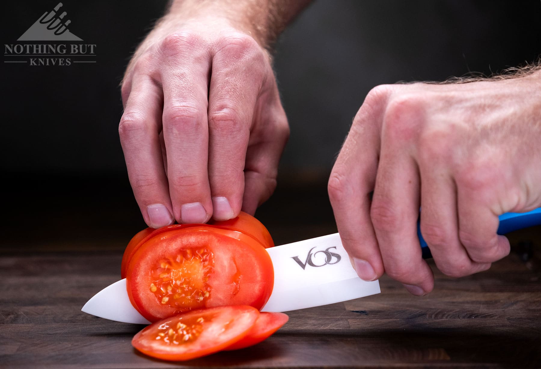 A close up of a tomatoe being sliced with a Vos ceramic chef knife on a wood cutting board in front of a dark background. 