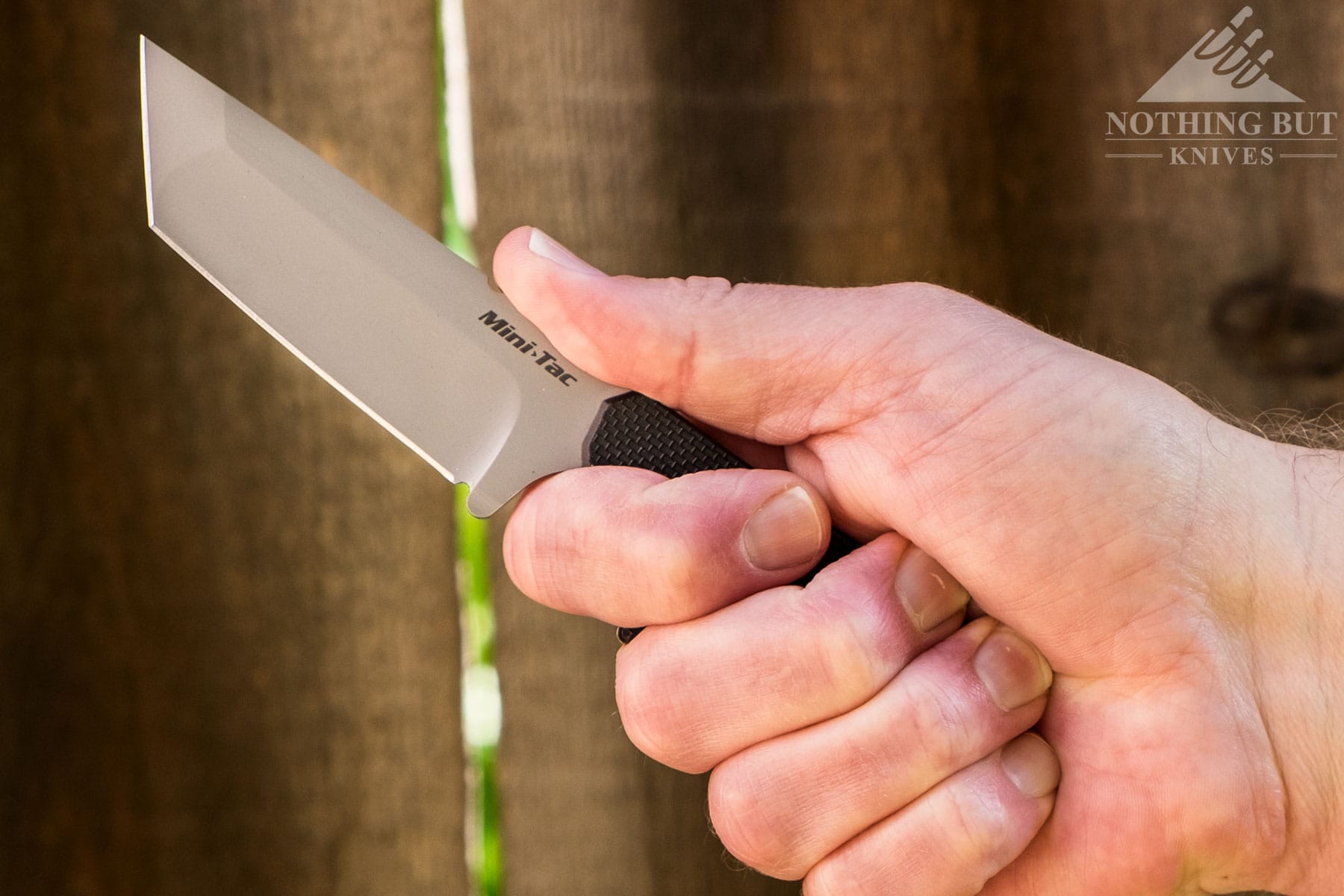 A close-up of a person's hand gripping the Mini Tac neck knife in front of a wooden fence. 