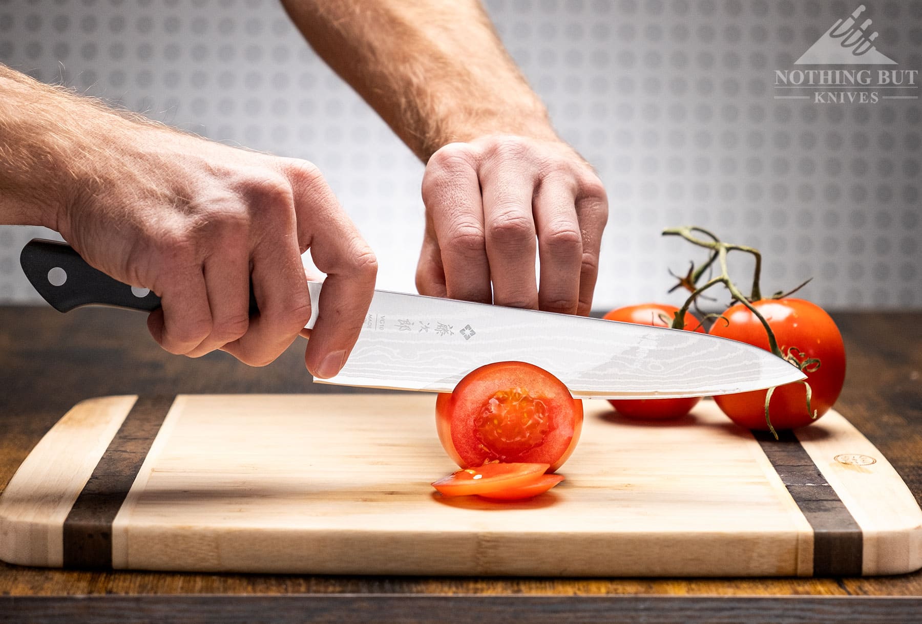 The Tojiro DP Damascus chef knife being used to slice a tomato on top of a bamboo cutting board in front of an off-white background. 