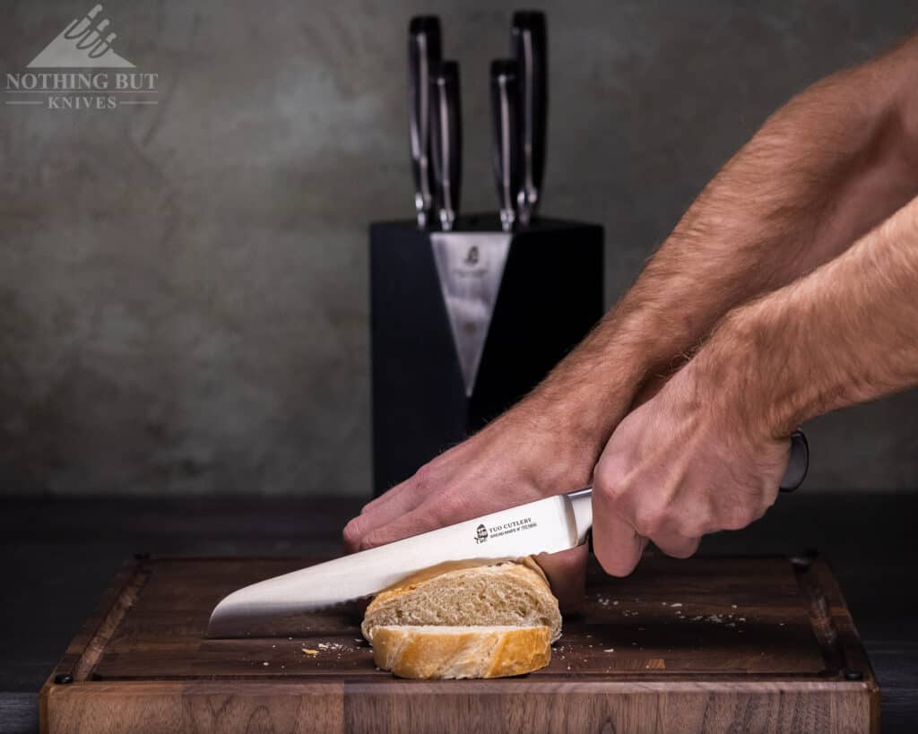 The Tuo 1810 bread knife being used to slice a loaf of French bread on an end grain cutting board next to the rest of the knife set.