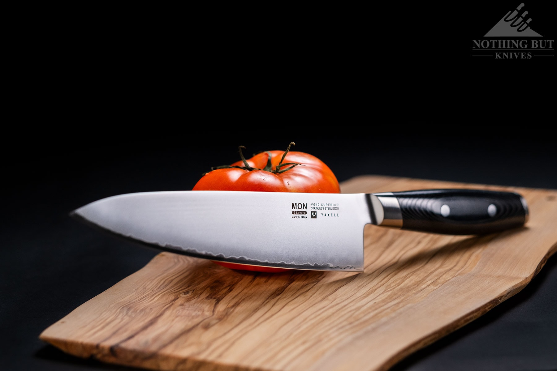 A Yaxell Mon Japanese chef knife leaning against a tomato on a wood cutting board.
