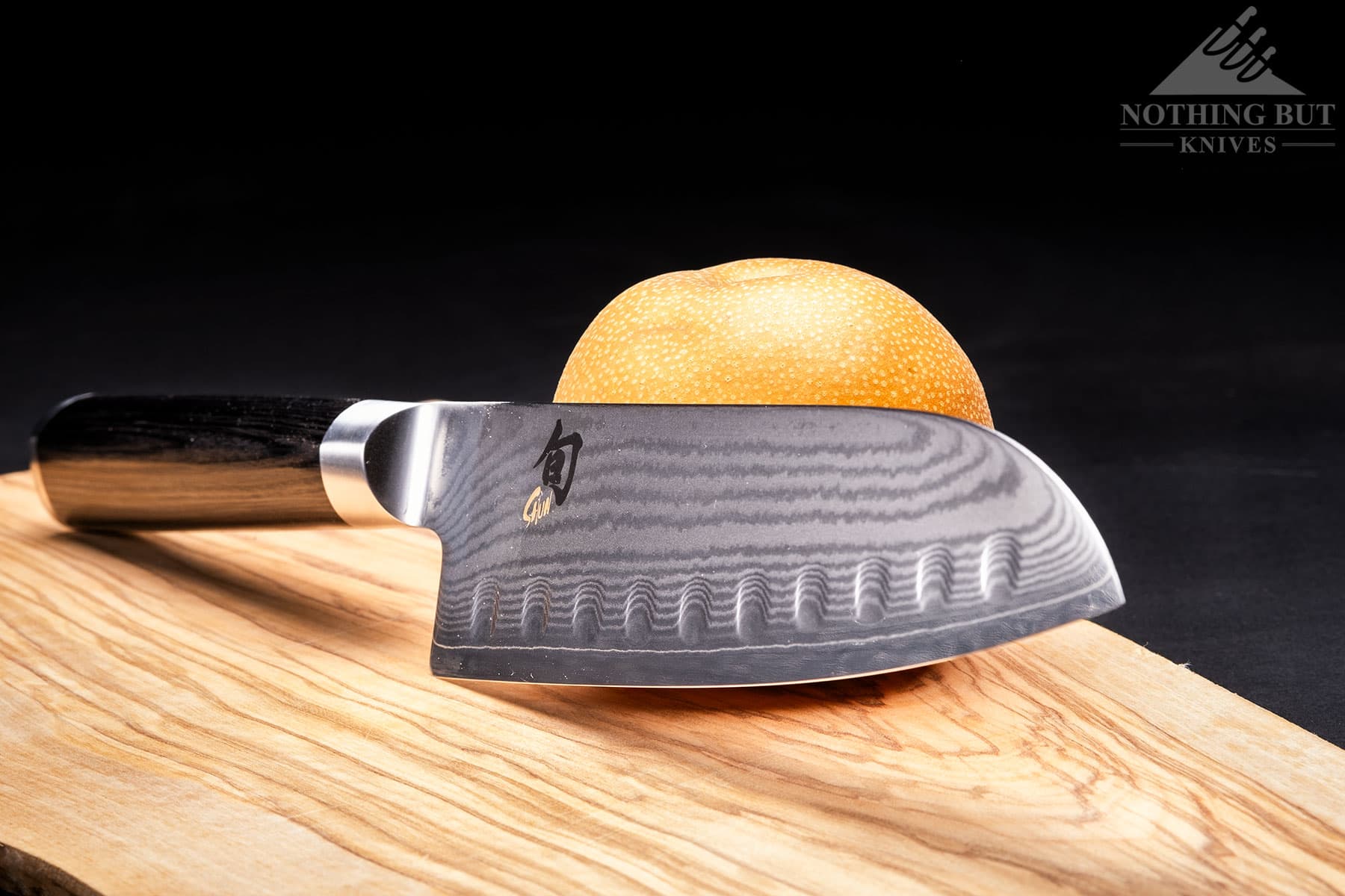 The Shun Classic Santoku knife leaning against an Asian pear on a wood cutting board.