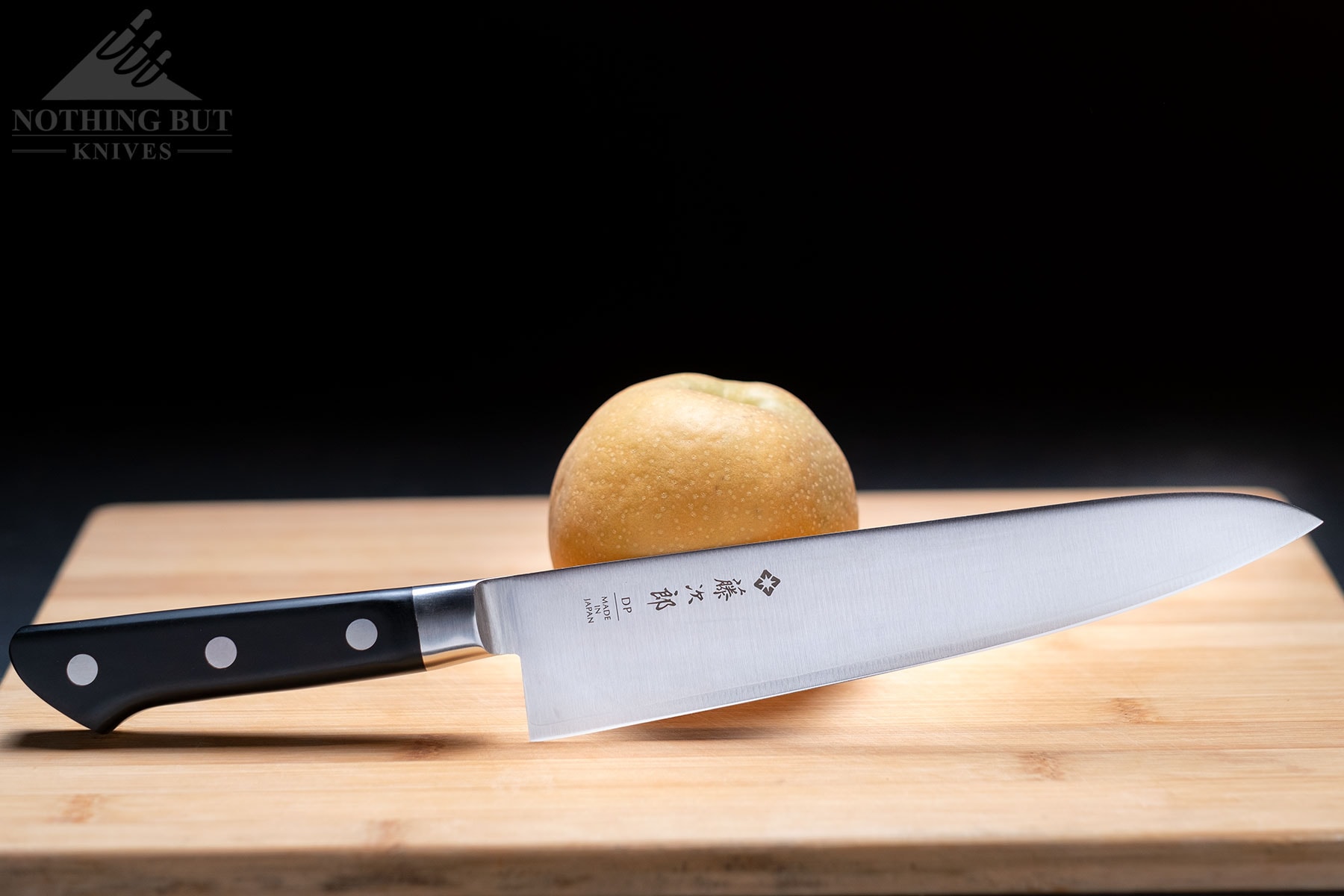 An 8-inch Tojiro DP chef knife leaning against a pear on a bamboo cutting board in front of a black background.