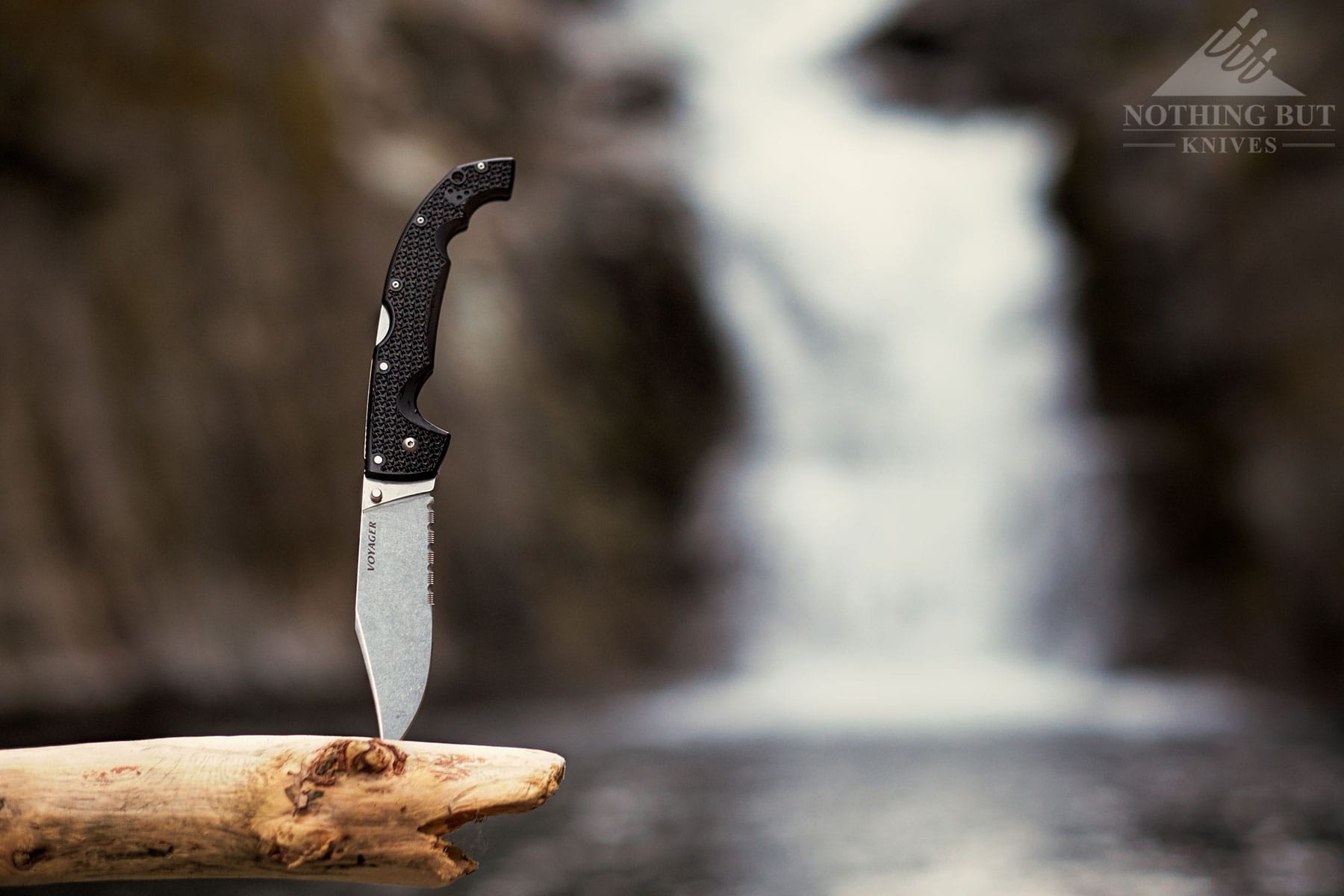 The Cold Steel Voyager pocket knife sticking out of a log in front of a waterfall.