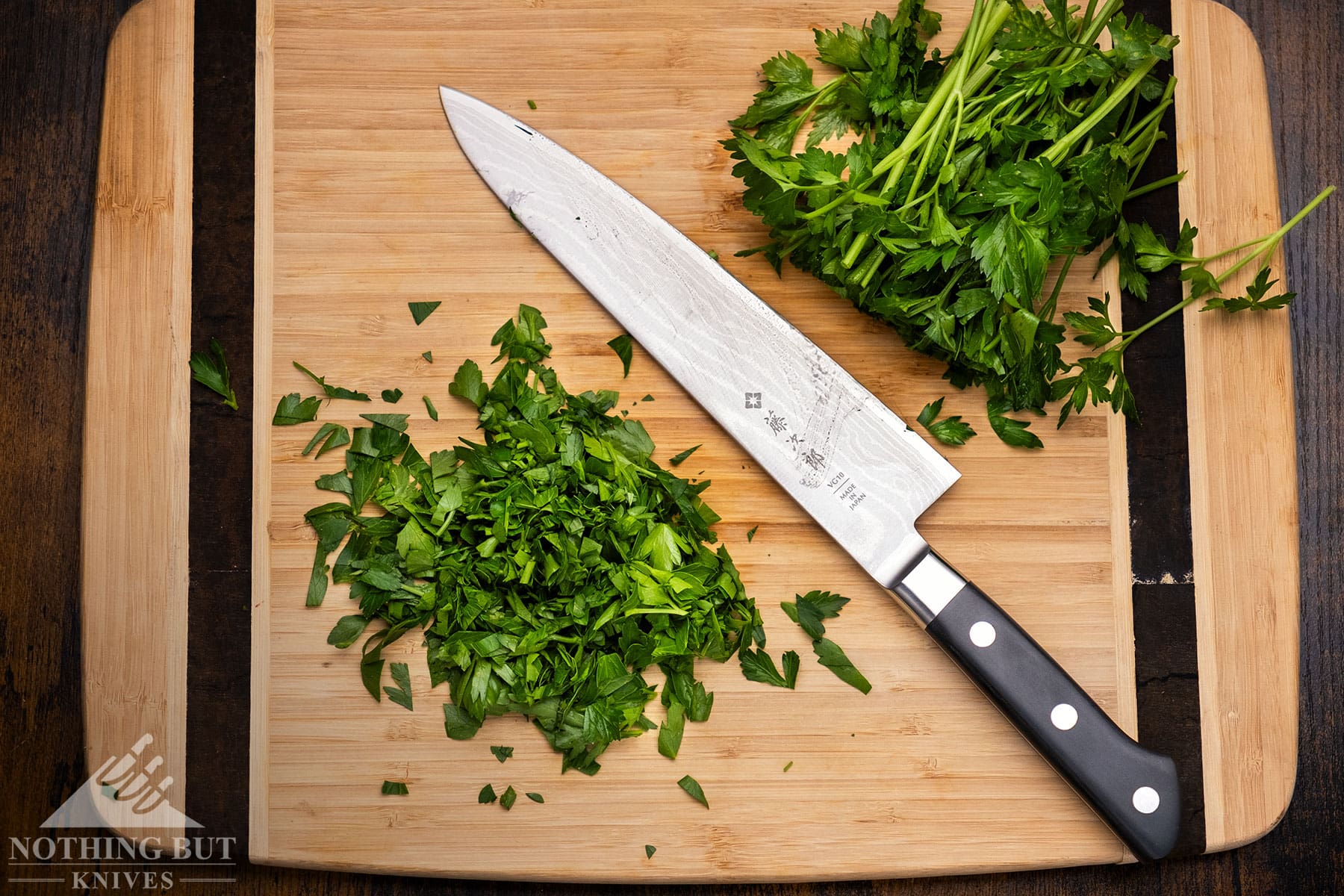 An overhead view of the Tojiro Dp Damascus chef knife between two piles of herbs it was used to chop on a bamboo cutting board. 