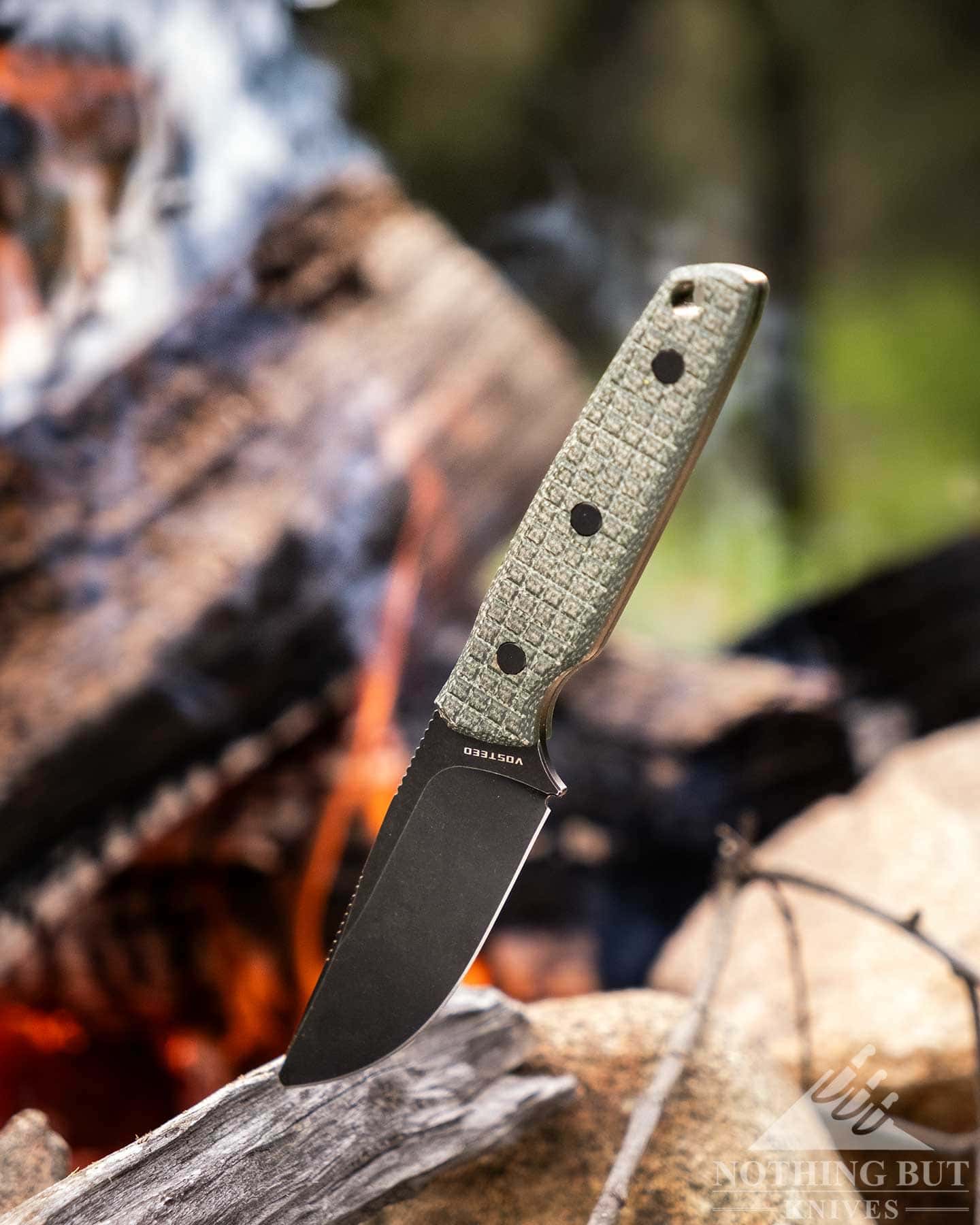 The Vosteed Mink fixed blade EDC knife shown here with its blade tip sticking out of a piece of wood in front of a campfire in a forest. 