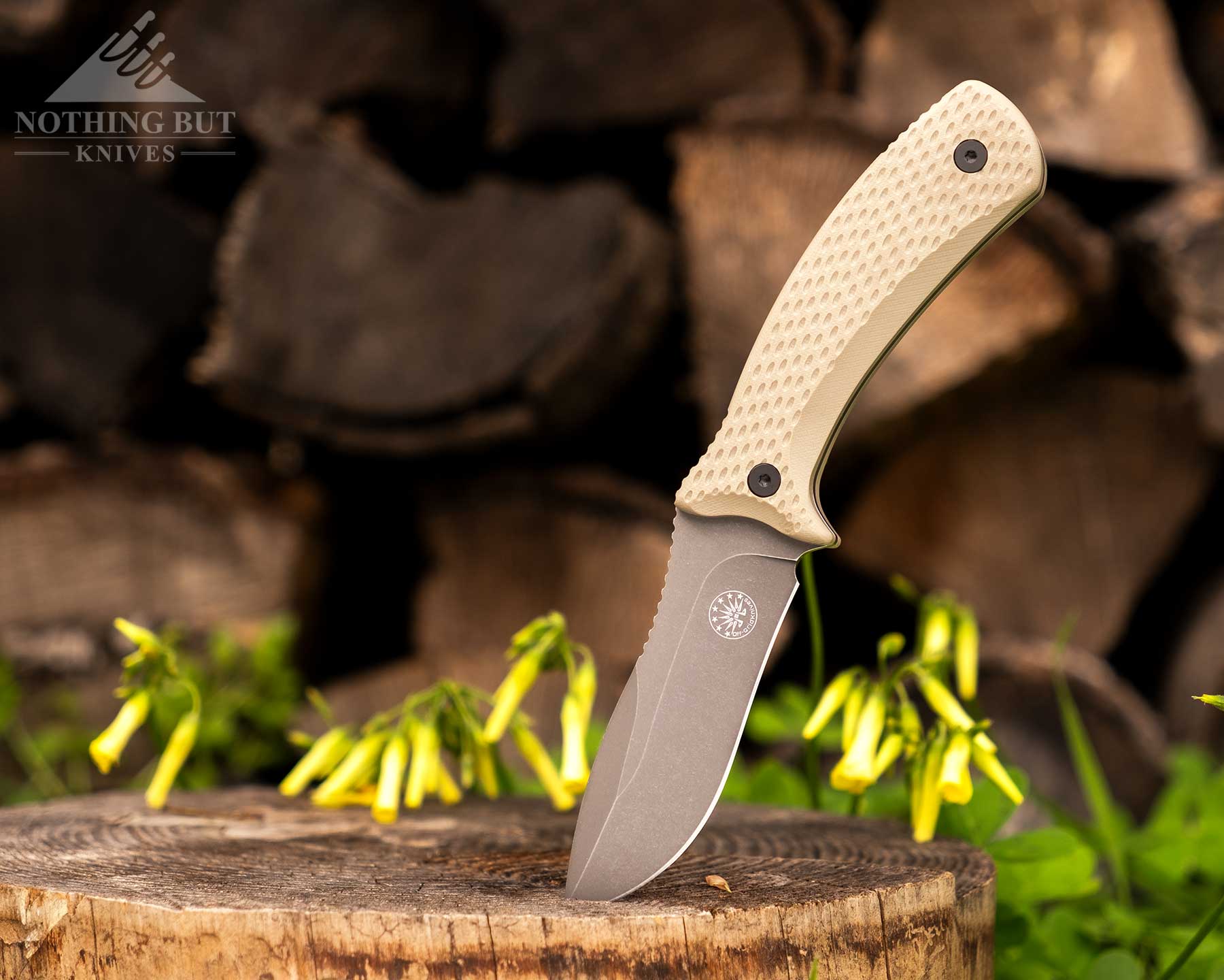 The Coyote Tan version of the Off-Grid Sidekick with its blade stuck in a log in front of a stack of firewood outdoors. 