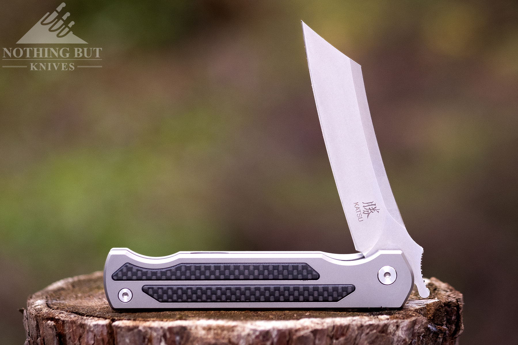 The Katsu Jt01 Camping knife shown here on a piece of firewood with the blade in the half open position in front of a blurry forest background.