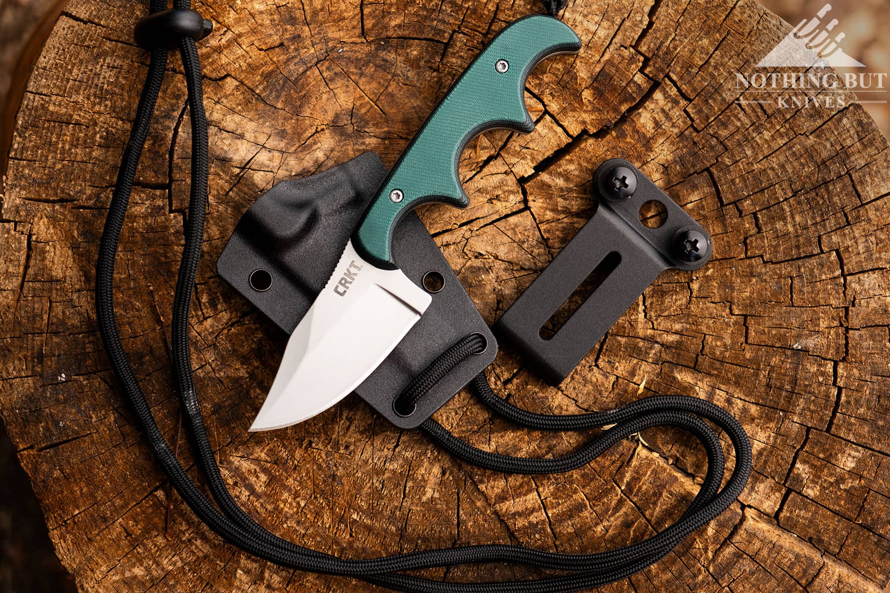 The CRKT Minimalist neck knife shown here on top of a tree stump with its sheath, lanyard and belt clip.