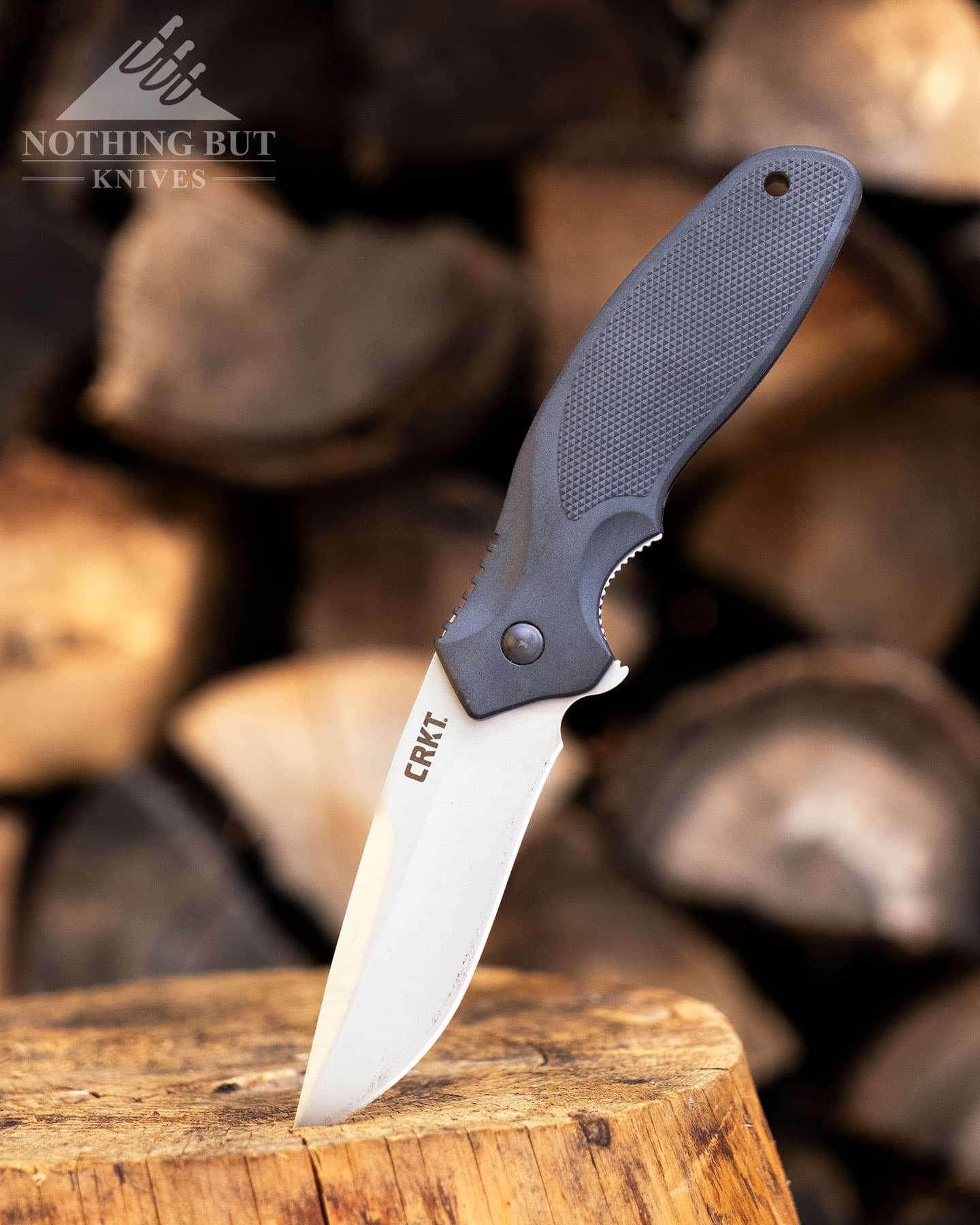 The CRKT Shenanagin Z budget friendly pocketknife shown here with the tip of the blade sticking out of a piece of firewood in front of stacked firewood. 