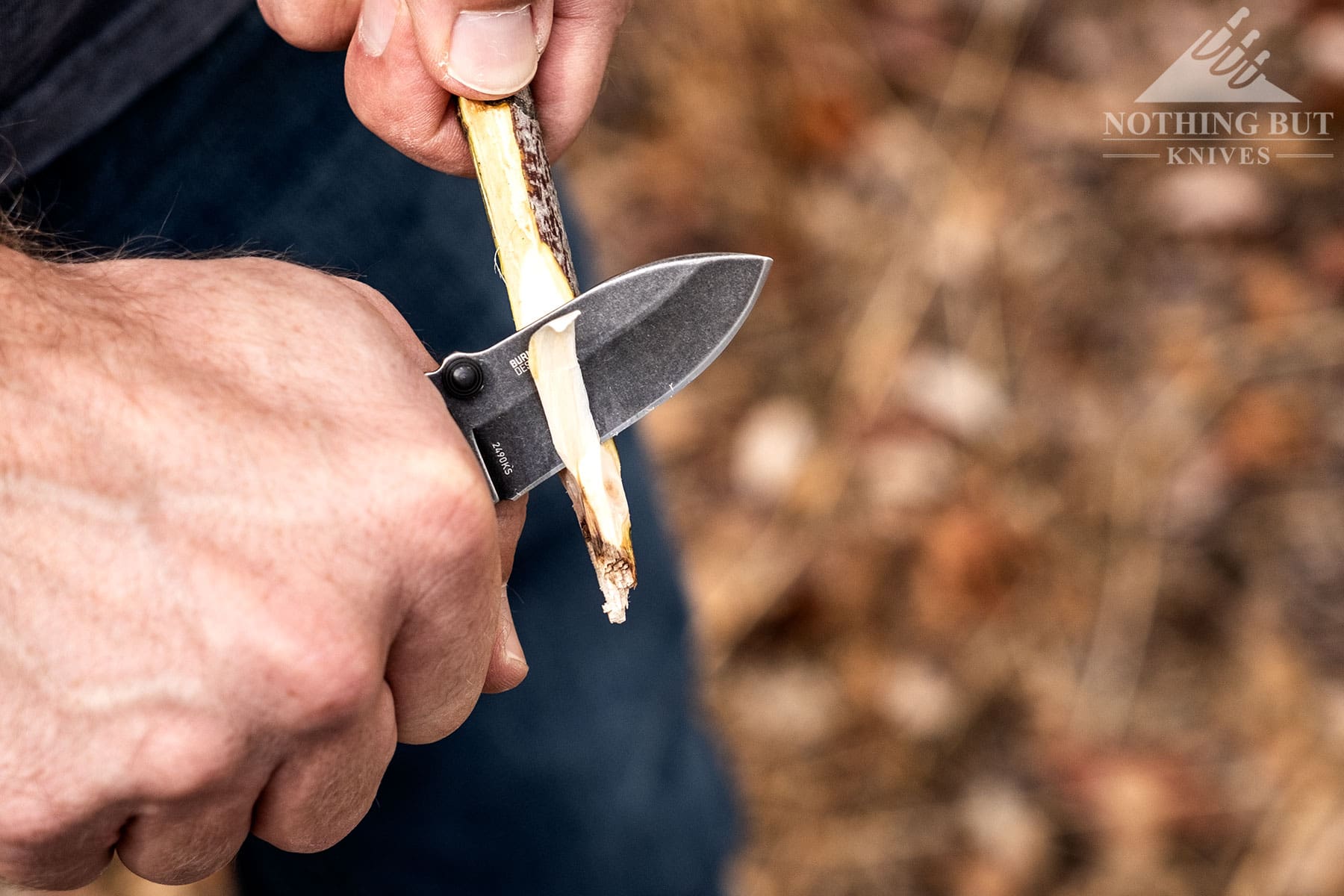 An overhead view of the CRKT Squid being used to carve a stick above a blurry leaf covered ground. 