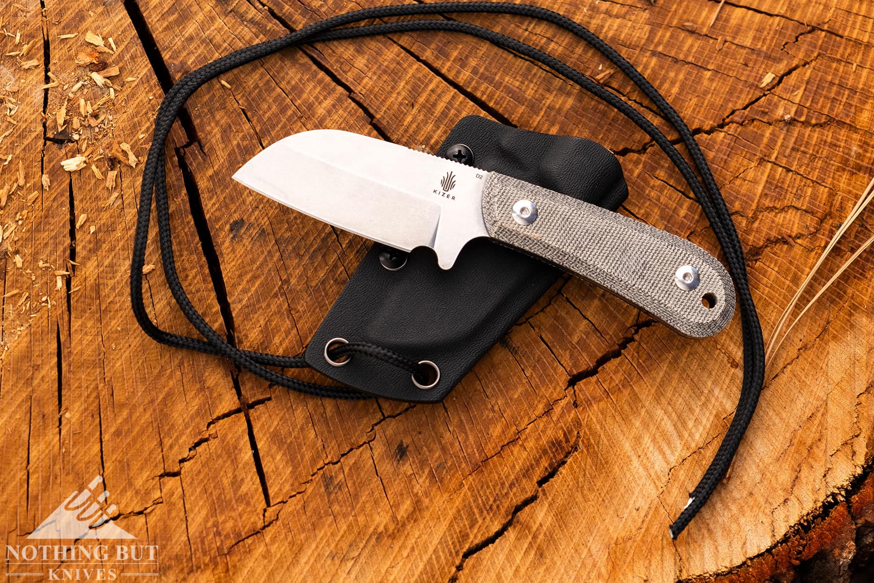 The EDC Deckhand D2 neck knife shown here with its kydex sheath and lanyard on top of a tree stump outdoors.