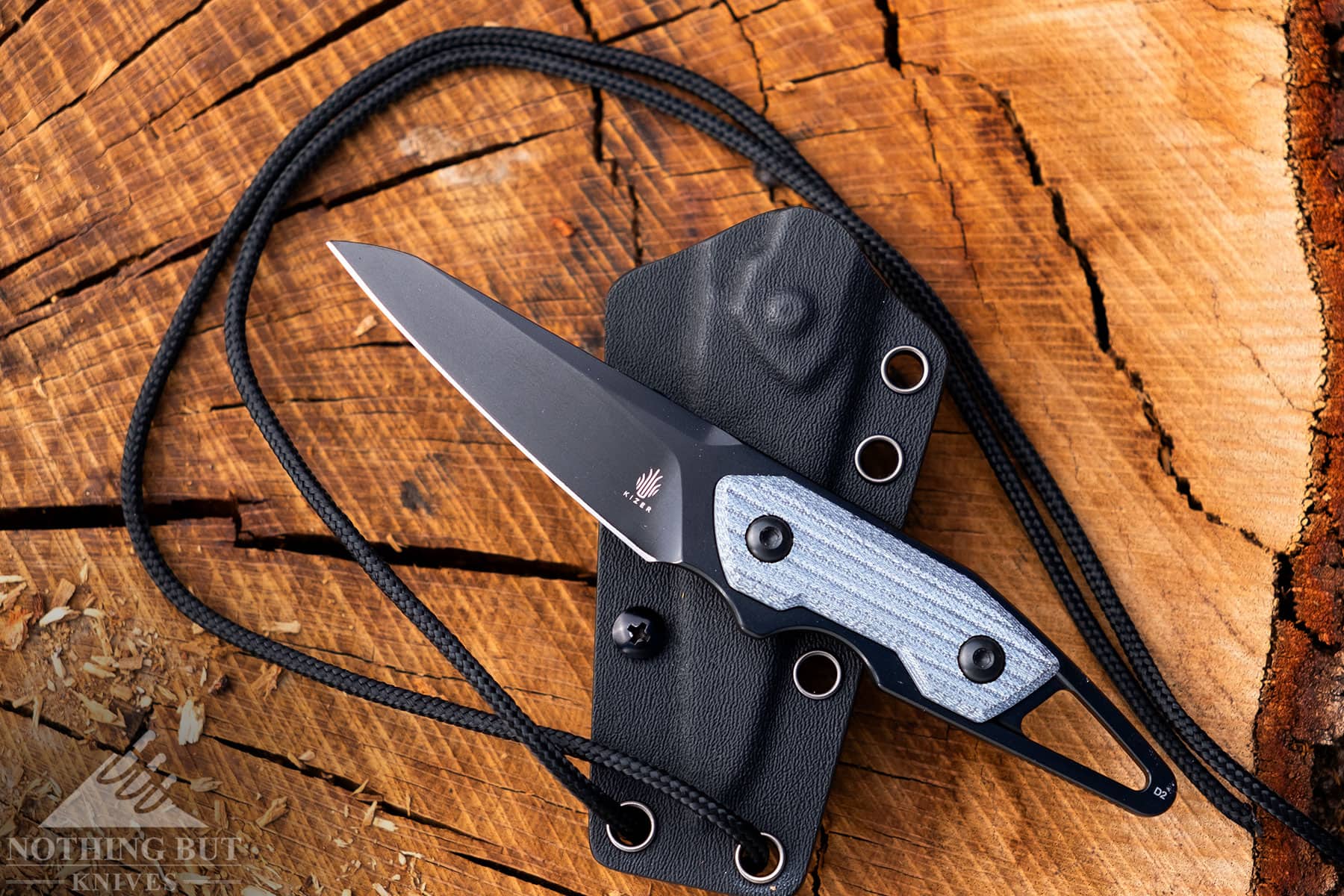 The Kizer fixed blade neck knife shown outside of its kydex sheath with its lanyard on top of a tree stump. 