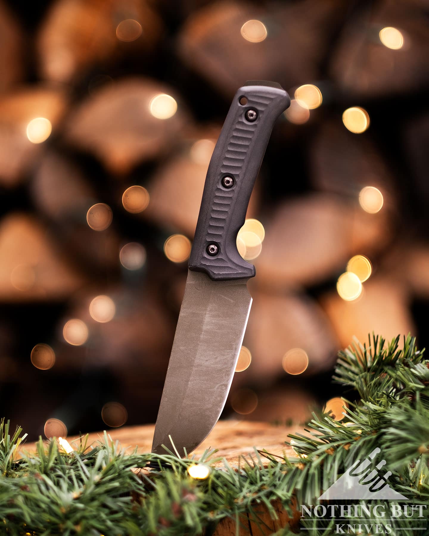 The Shwarz Lost Trail 5 camp knife with its blade stuck in a tree stump in front of a woodpile covered in Christmas lights.