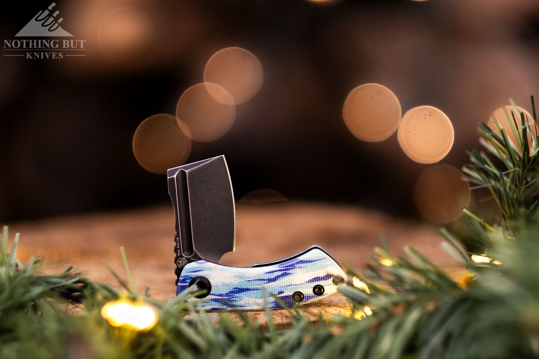 The Ice version of the tiny Kansept Mini Korvid pocket knife in the half open position on a tree stump decorated with lights for the holidays.