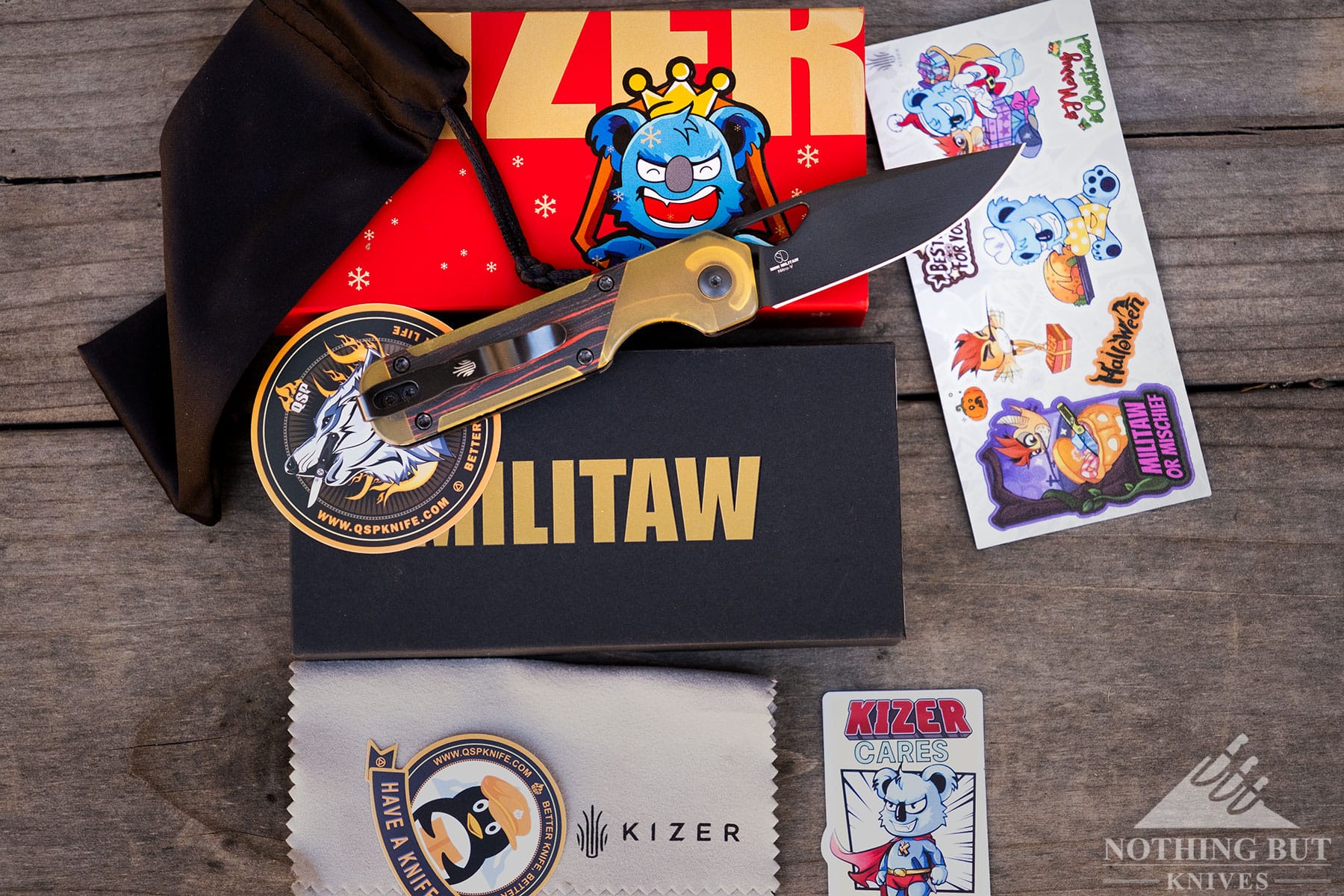 An overhead view of the Kizer Mini Militaw with its blade open on top of its packaging surrounded by the swag that is included. 