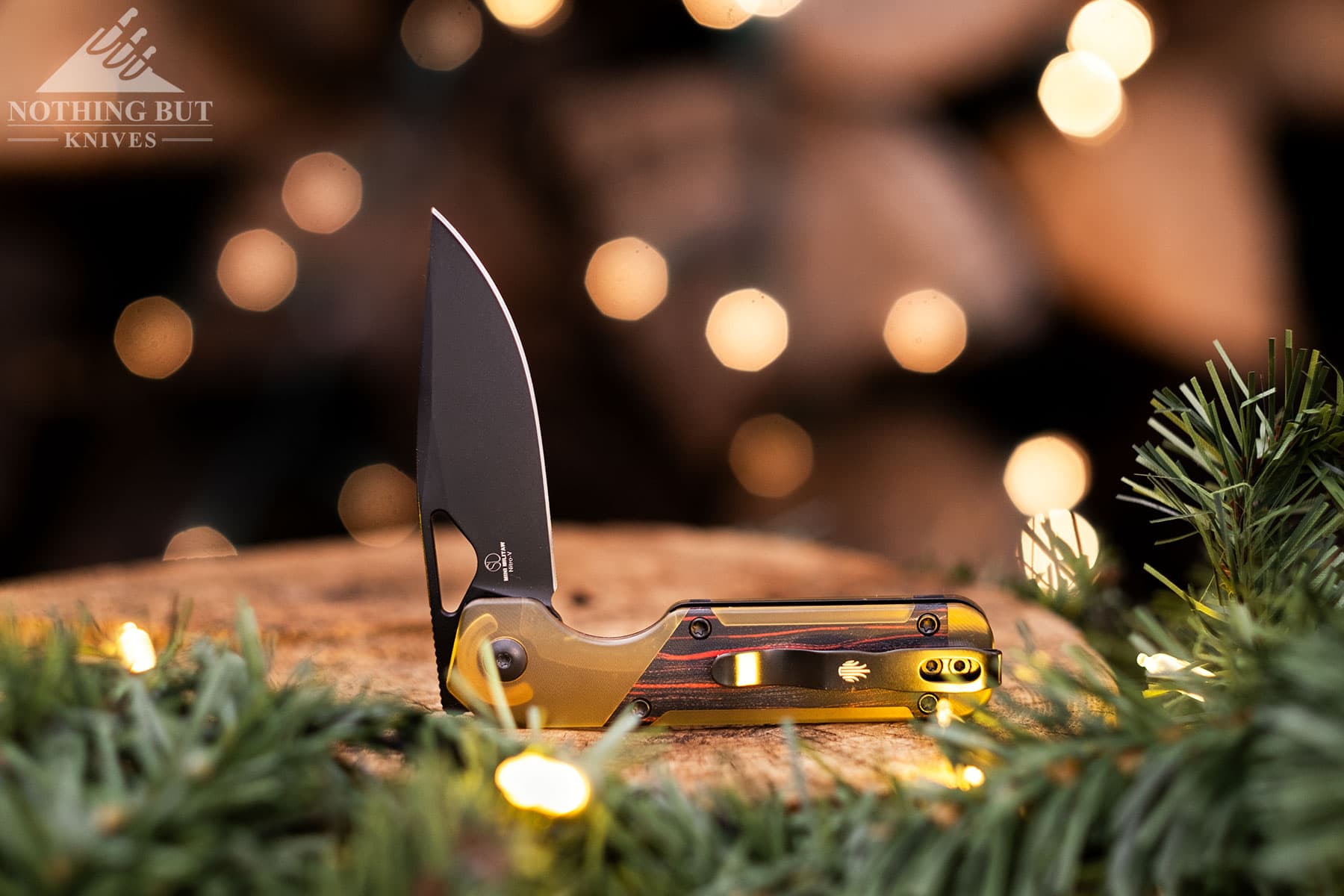 The Kizer Militaw Mini in the half open position in front of a stack of firewood covered in Christmas lights. 