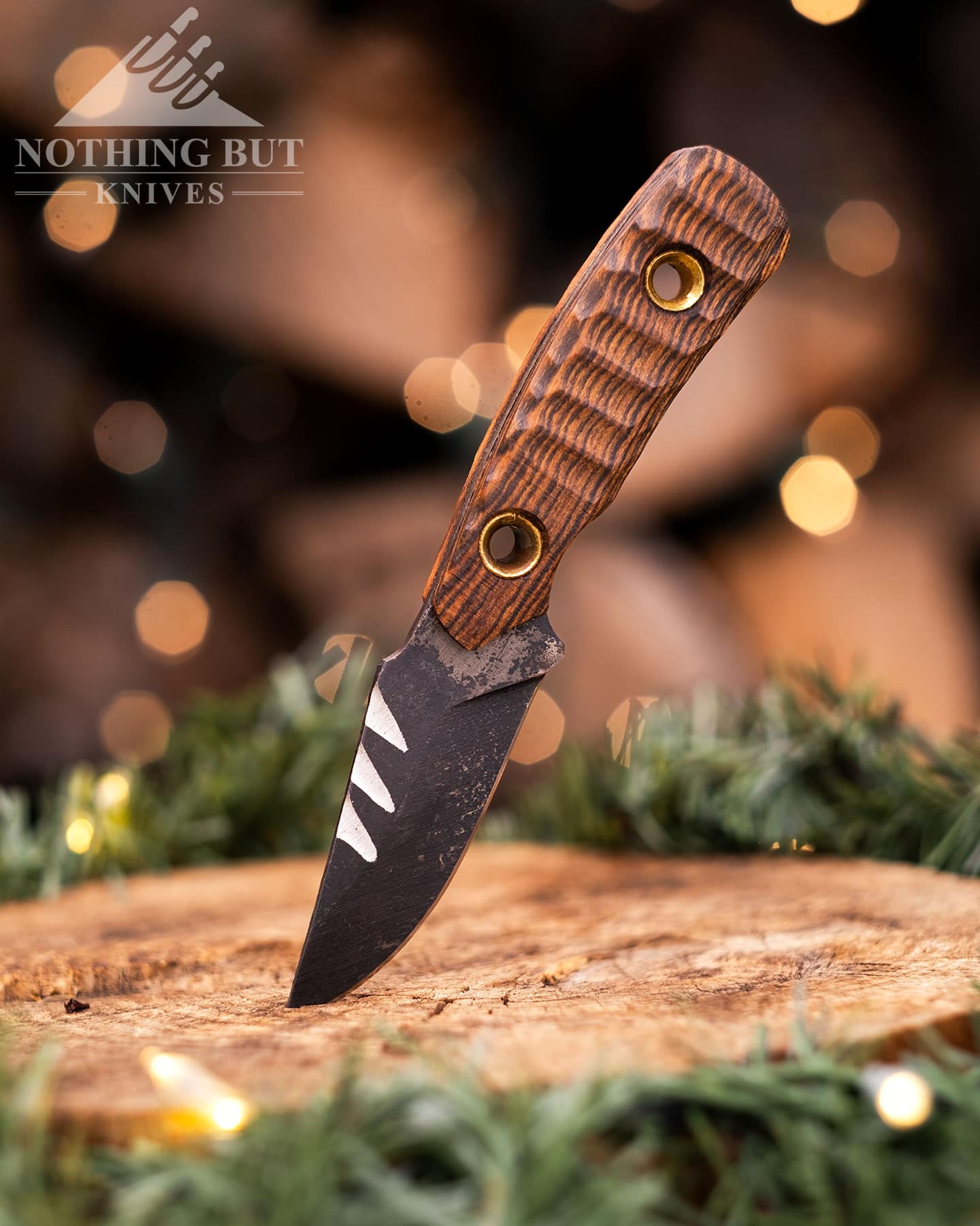 The Texas Cowboy knife sticking out of a piece of firwood in front of a stack of firewood decorated with holiday lights. 