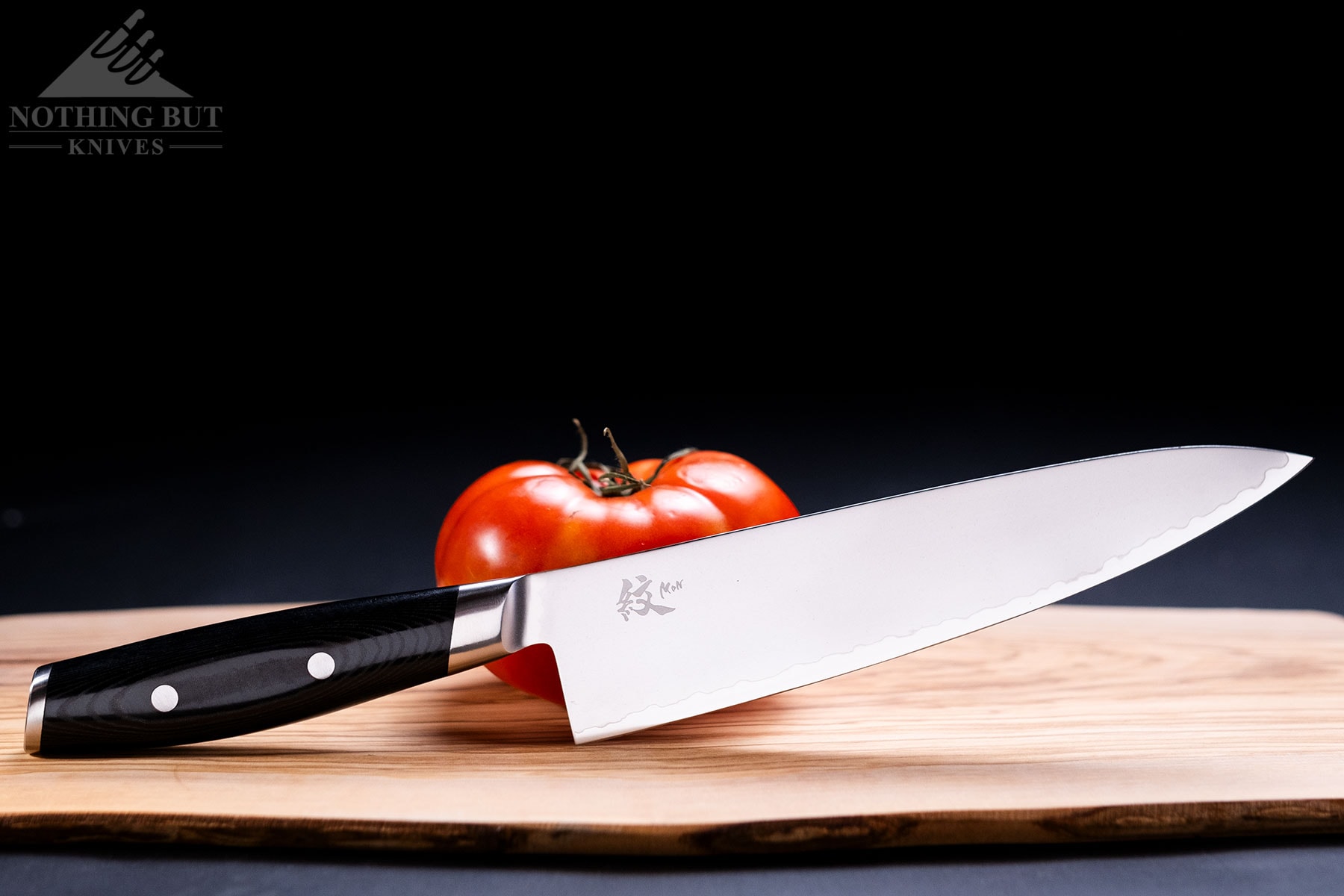 A Yaxell Mon 8-inch chef knife on a wood cutting board with a tomato in front of a black background.