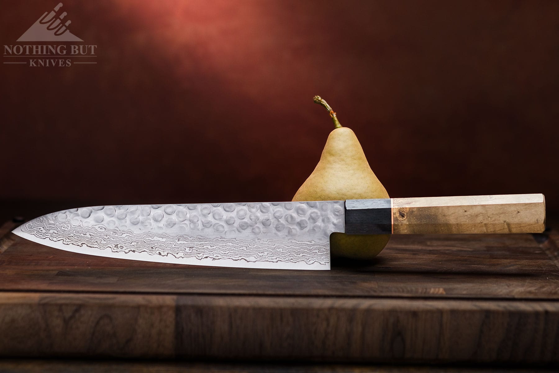 A modern Damascus steel kitchen knife next to a pear on a dark wood cutting board. 