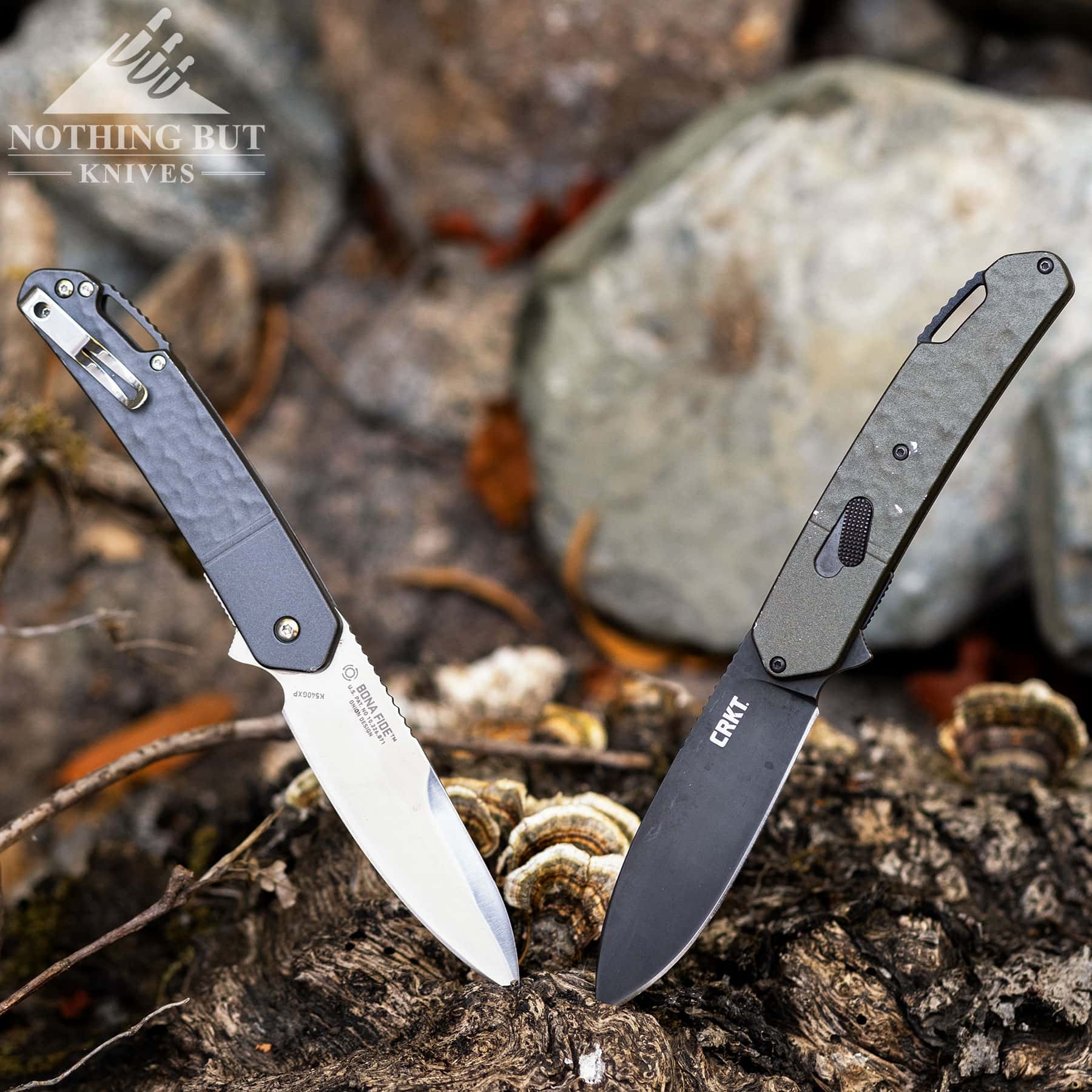 The two versions of the CRKT folding knife sticking out of a tree stump in front of a pile of rocks. 