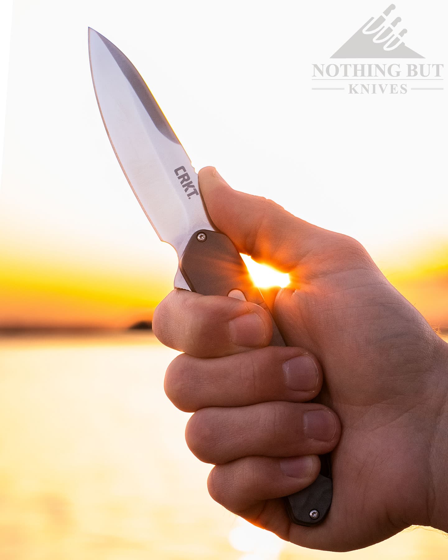 The silver version of the CRKT Bona Fide being gripped in a person's hand in front of a lake at sunset.