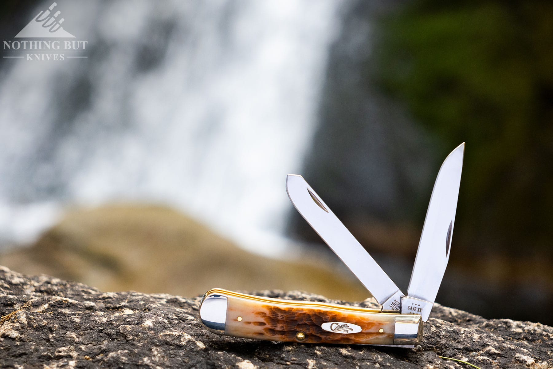 The Amber Bone version of the Case Knives Trapper slip joint with both blades open on a rock in front of a waterfall. 