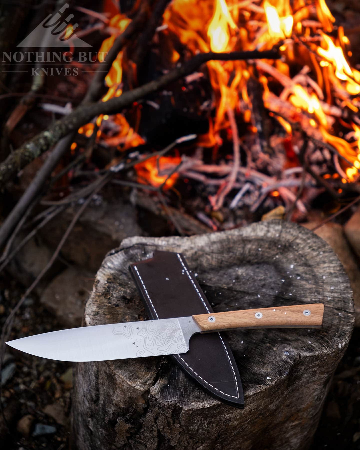 An overhead view of the BPS Rosemary camping chef knife on top of its leather sheath in front of a campfire. 