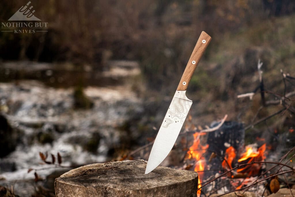 The BPS Rosemary camp cooking knife sticking out of a tree stump next to a campfire in front of a mountain creek. 