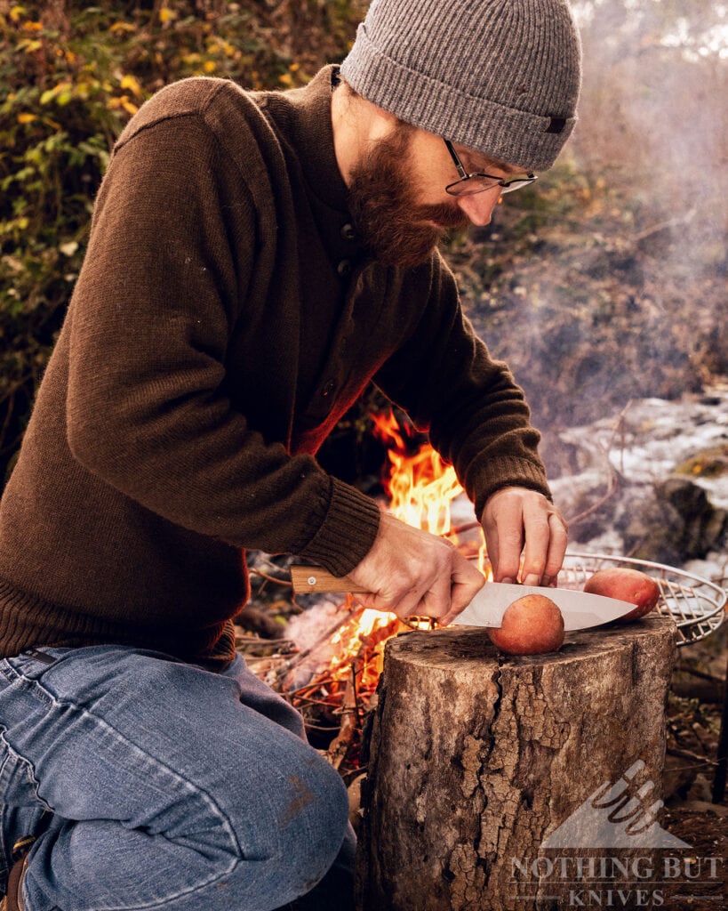 A man in a sweater and stocking cap kneeling by a campfire next to a creek using a camping chef knife to slice meat. 