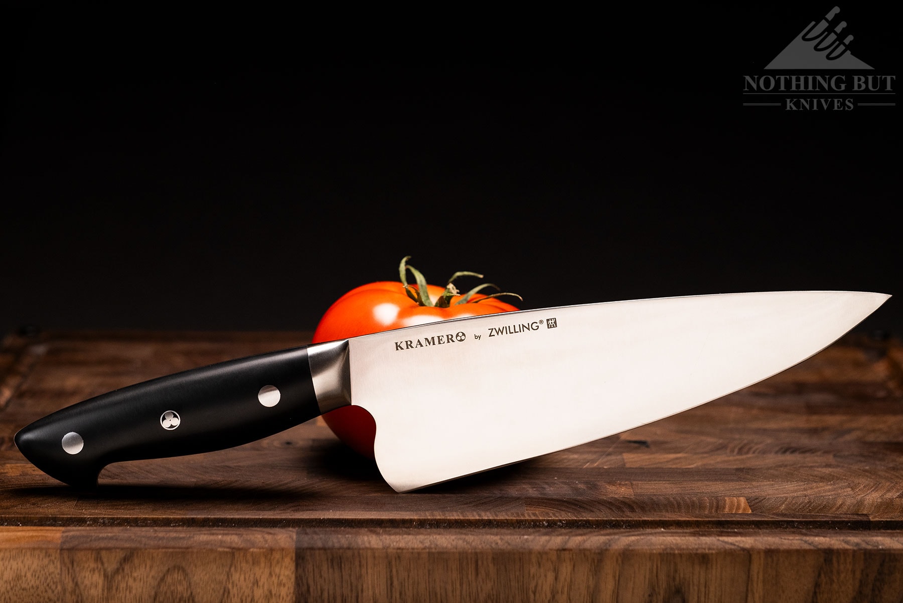 The Kramer By Zwilling Essentials Collection chef knife leaning against a ripe tomato on a wood cutting board.