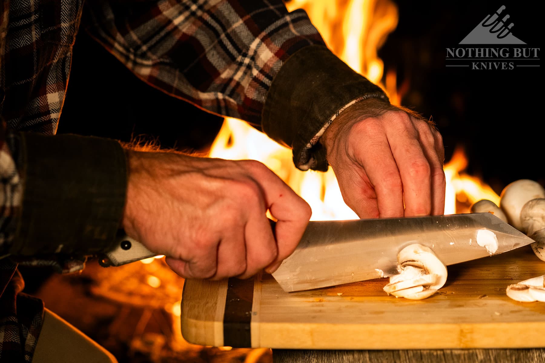 A close-up of the Messermeister Overland being used to slice mushrooms on a wood cutting board in front of a campfire. 