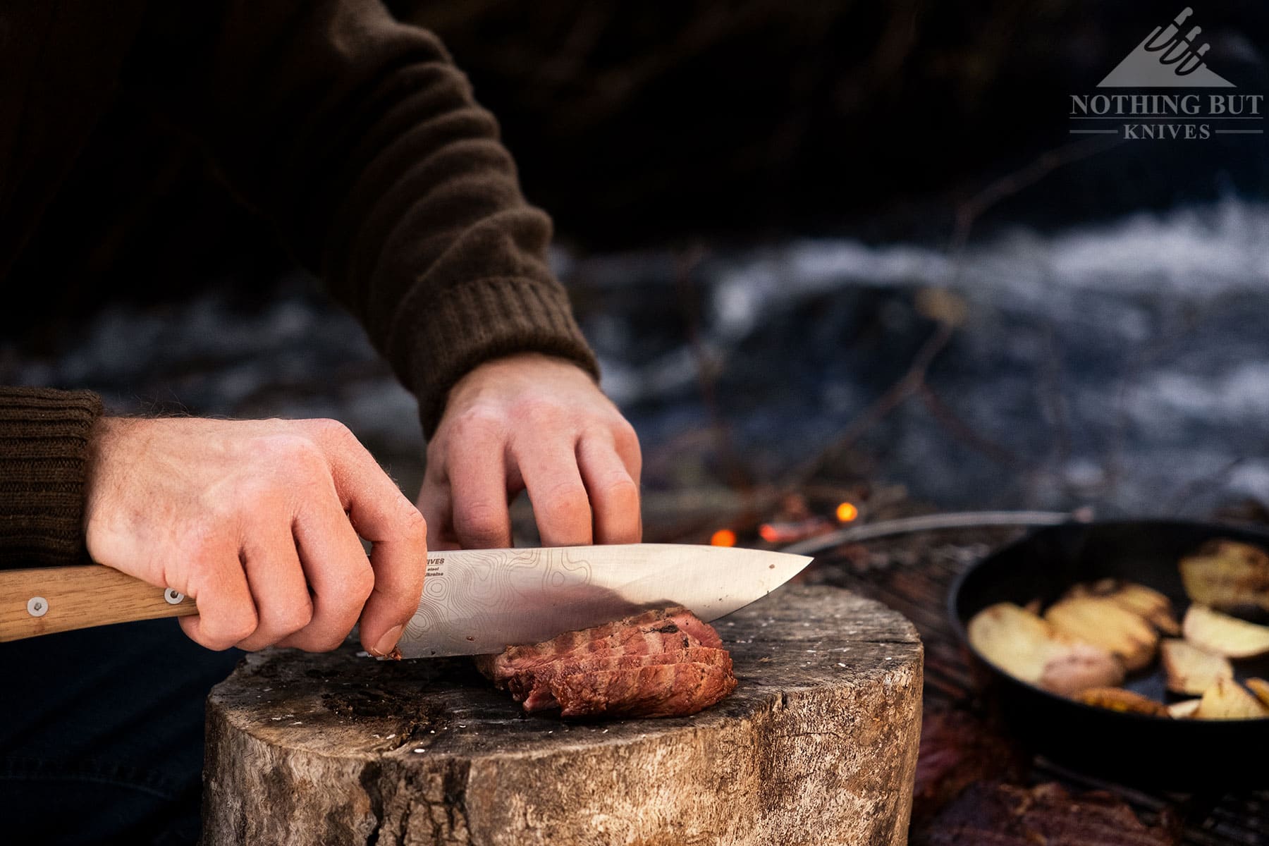 A close-up of the BPS Rosemary camp knife being used to slice steak next to a campfire with a skillet on the grill. 