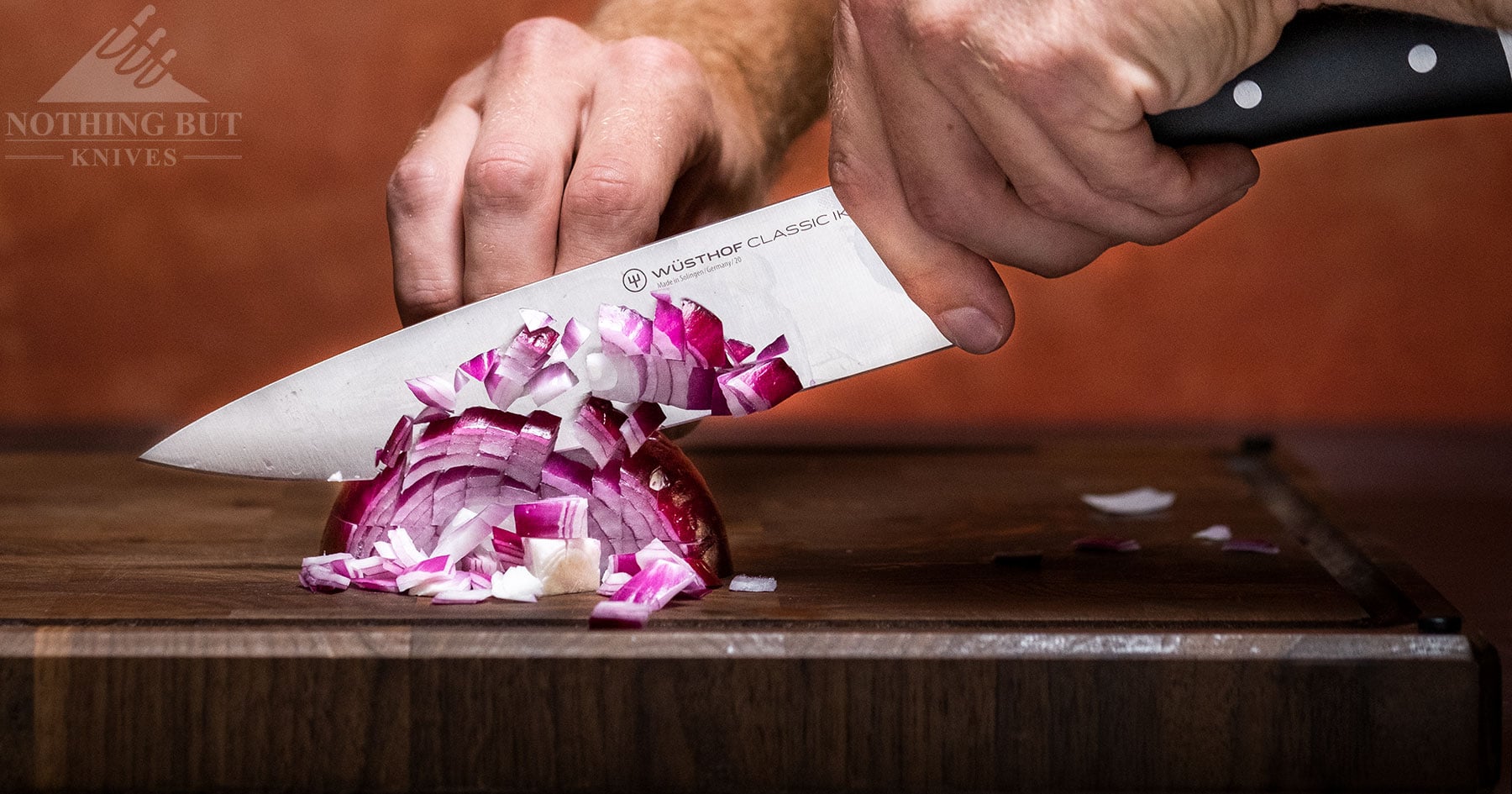 The Wusthof Classic Ikon shown here in the lefthand of a person using it to dice a red onion in front of a maroon background. 