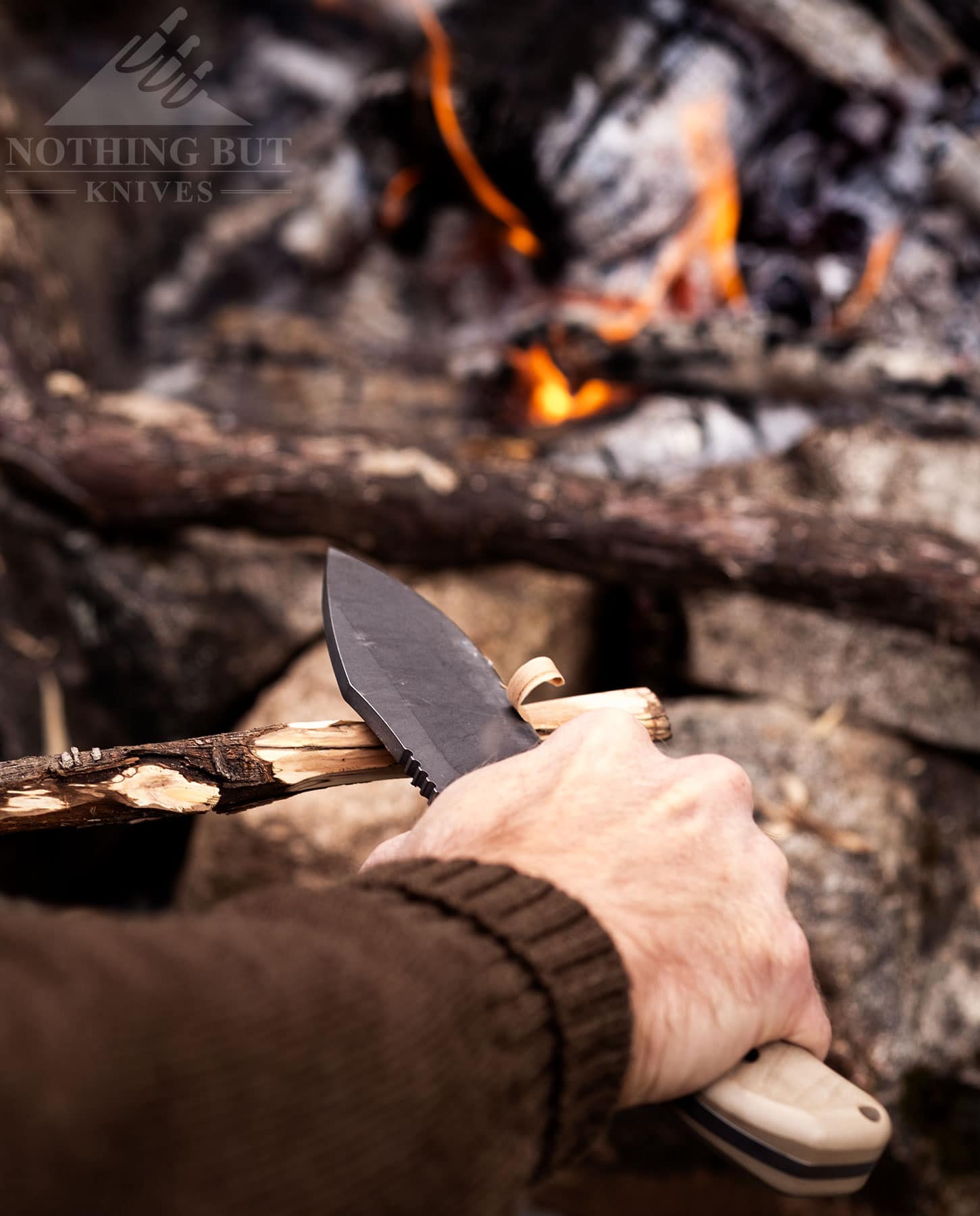 An overhead shot of a person's right hand using a survival knife to carve a stick by a campfire.