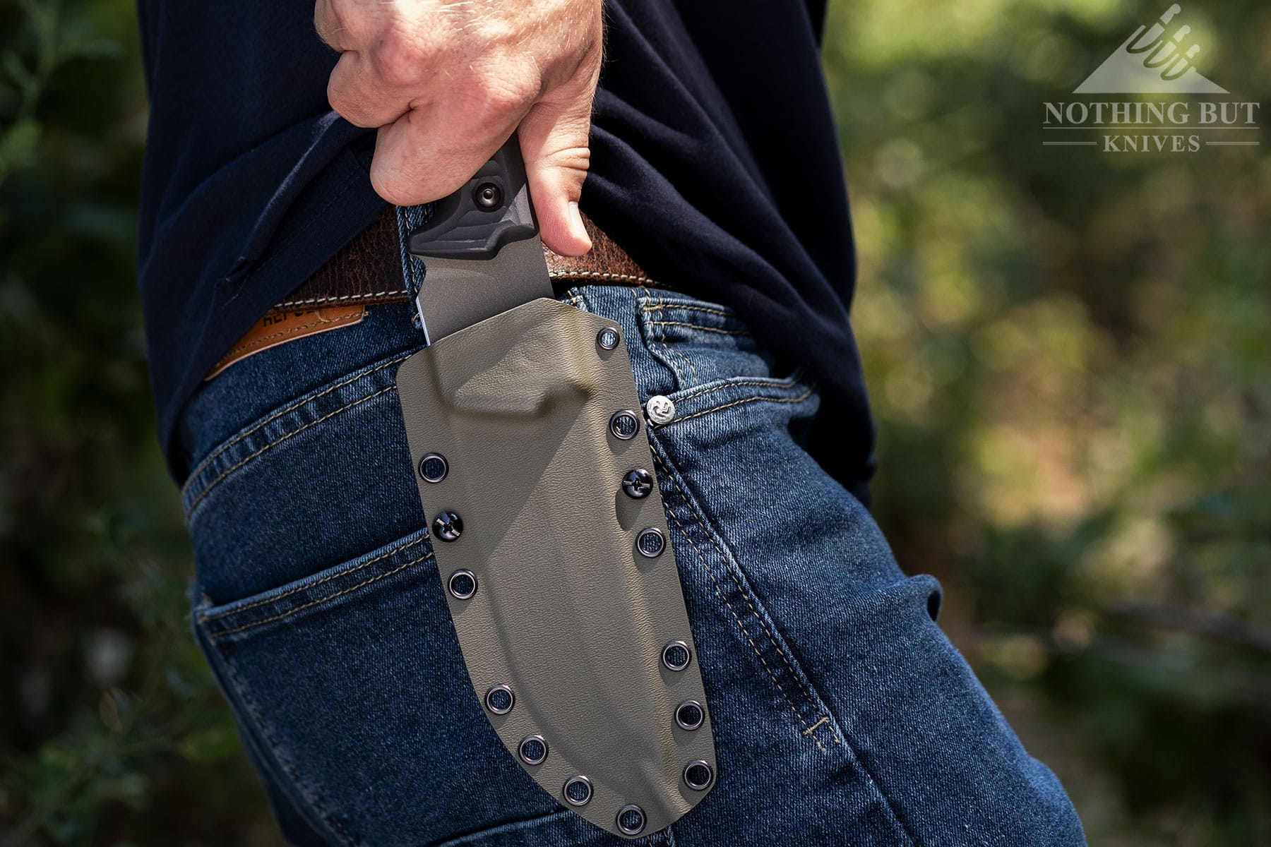 The Lost Trail 5 survival knife being deployed from it sheath by a person's right hand in front of a blurry background of trees. 