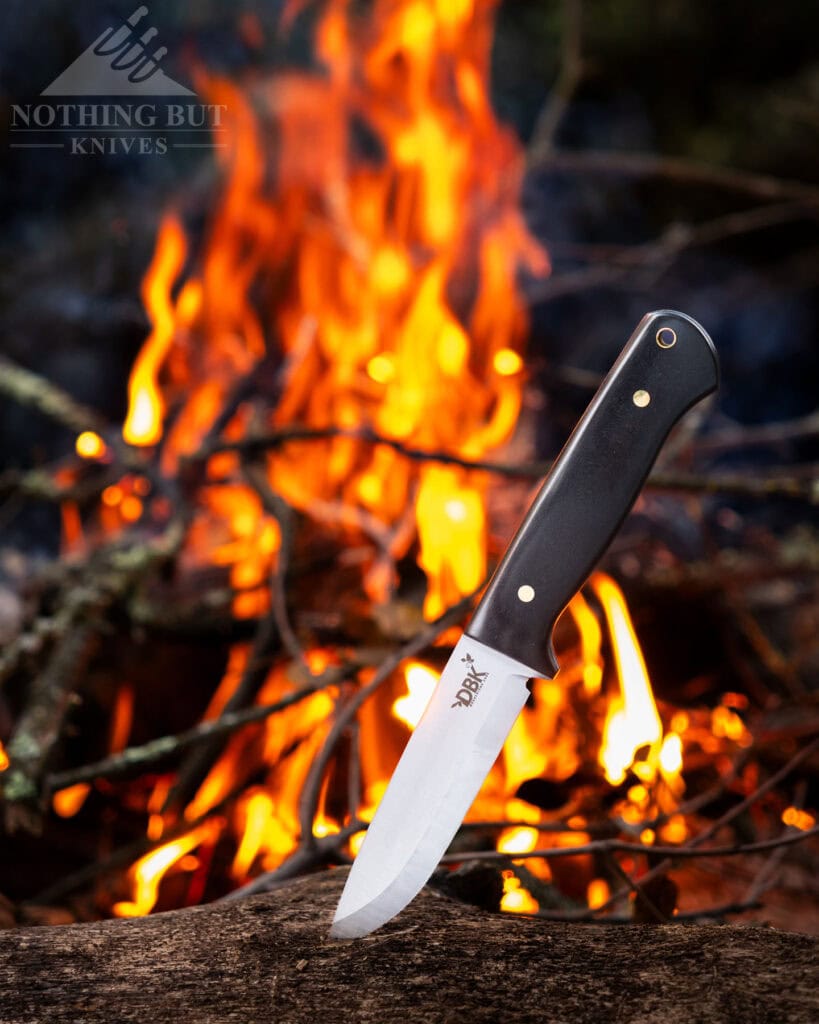 The Scandi grind version of the DBK Bushfriend 2000 with its blade tip sticking out of a piece of firewood in front of a campfire. 