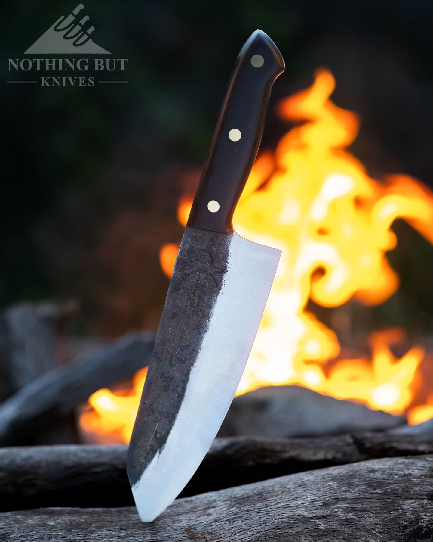 The Kitchen Perfection chef knife sticking out of a piece of firewood in front of a campfire.