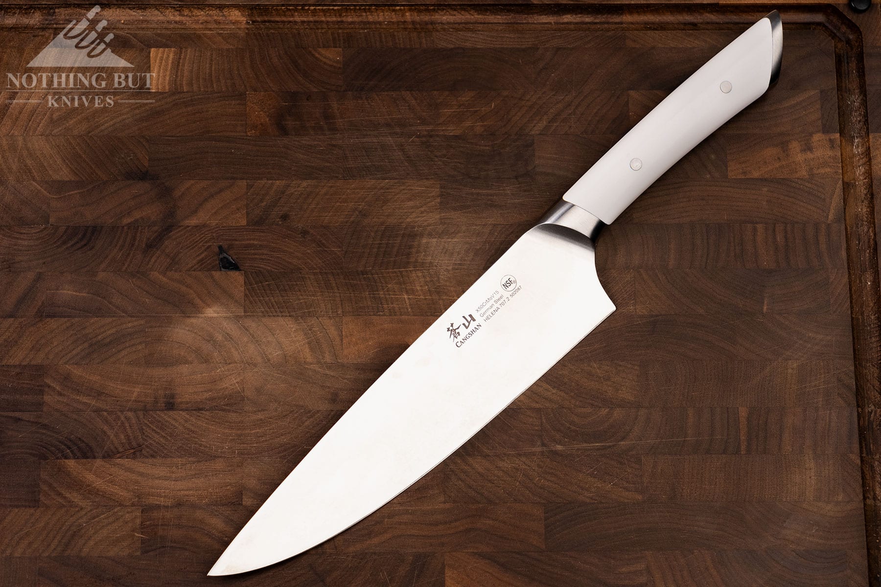 The Cangshan Helena eight inch chef knife with a white handle on a dark wood cutting board. 