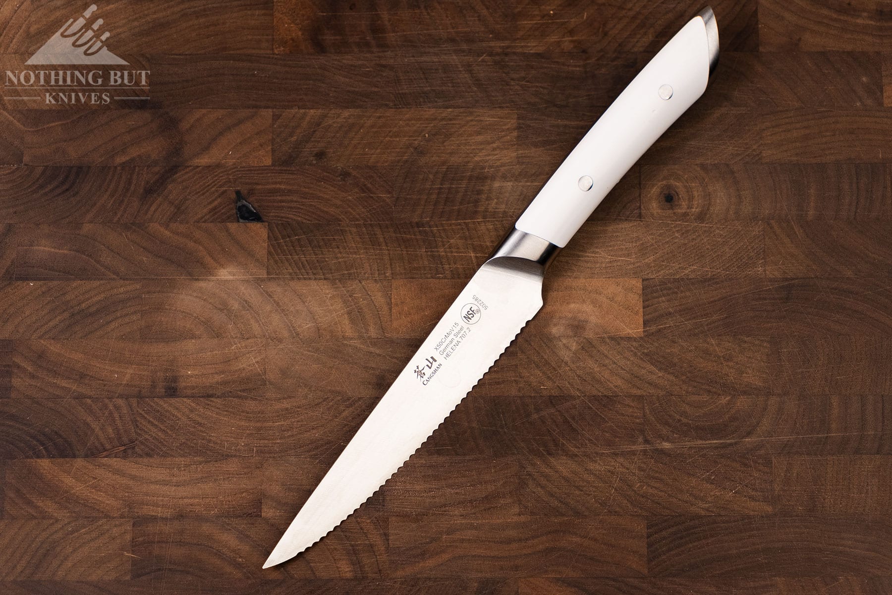 The Cangshan Helena serrated utility knife on a dark wood cutting board.