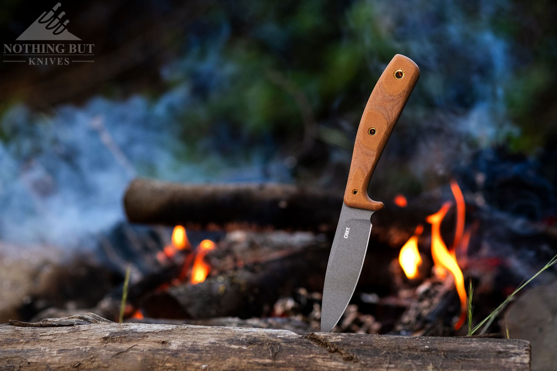 The CRKT Soldotna fixed blade hunting knife sticking out of a log in front of a campfire.