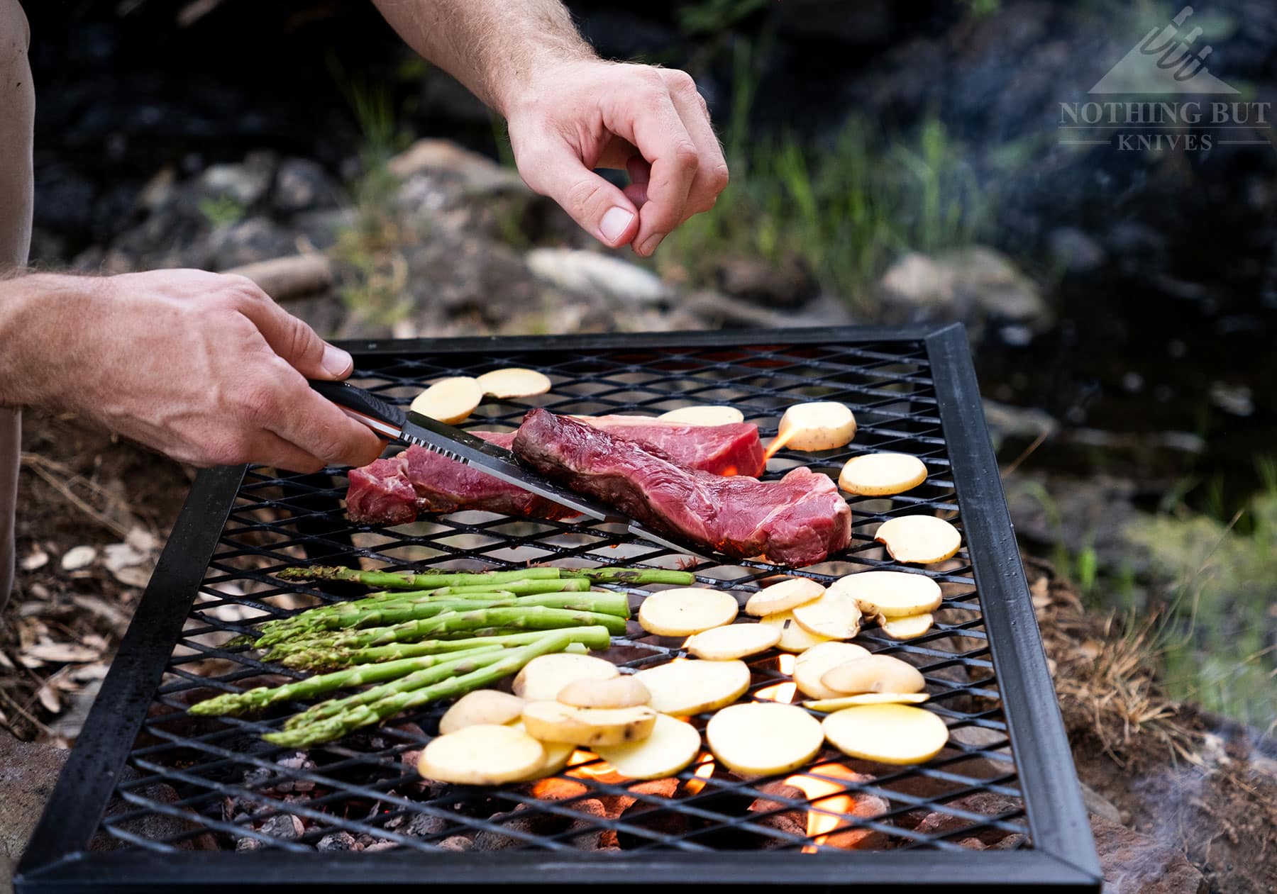 A close-up of the Maxsa Blade fixed blade knife being used to flip a steak on a campfire grill. 