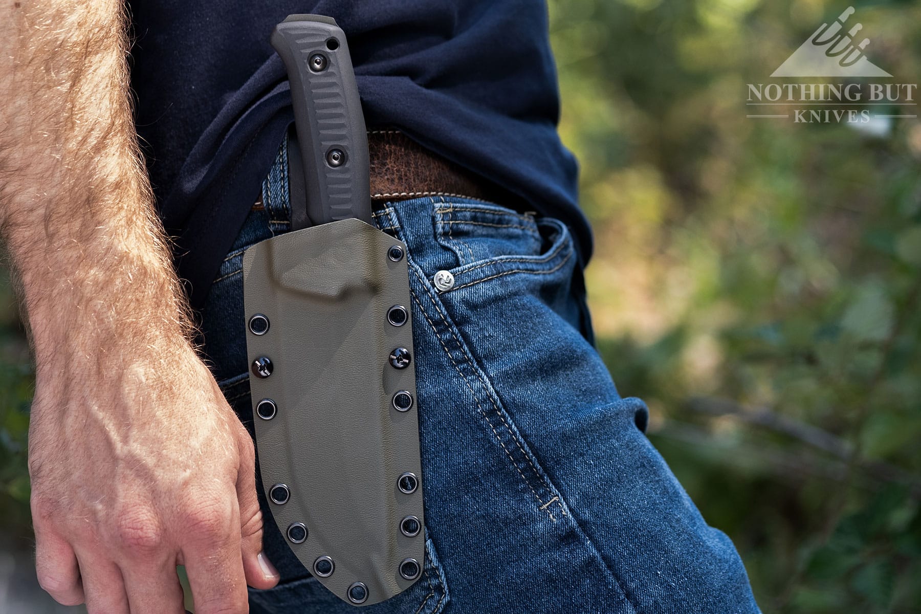 A close-up of the Lost Trail 5 fixed blade knife in its sheath hanging from a belt on a person's waist.