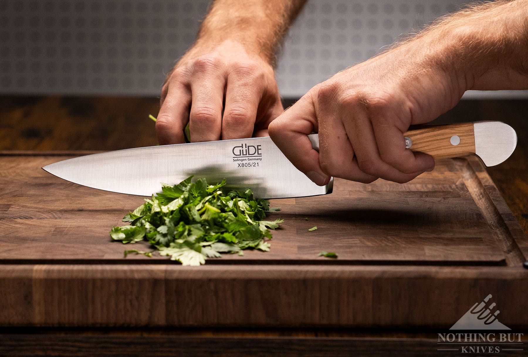 A Gude 8-inch chef knife being used to dice herbs on a wood cutting board. 