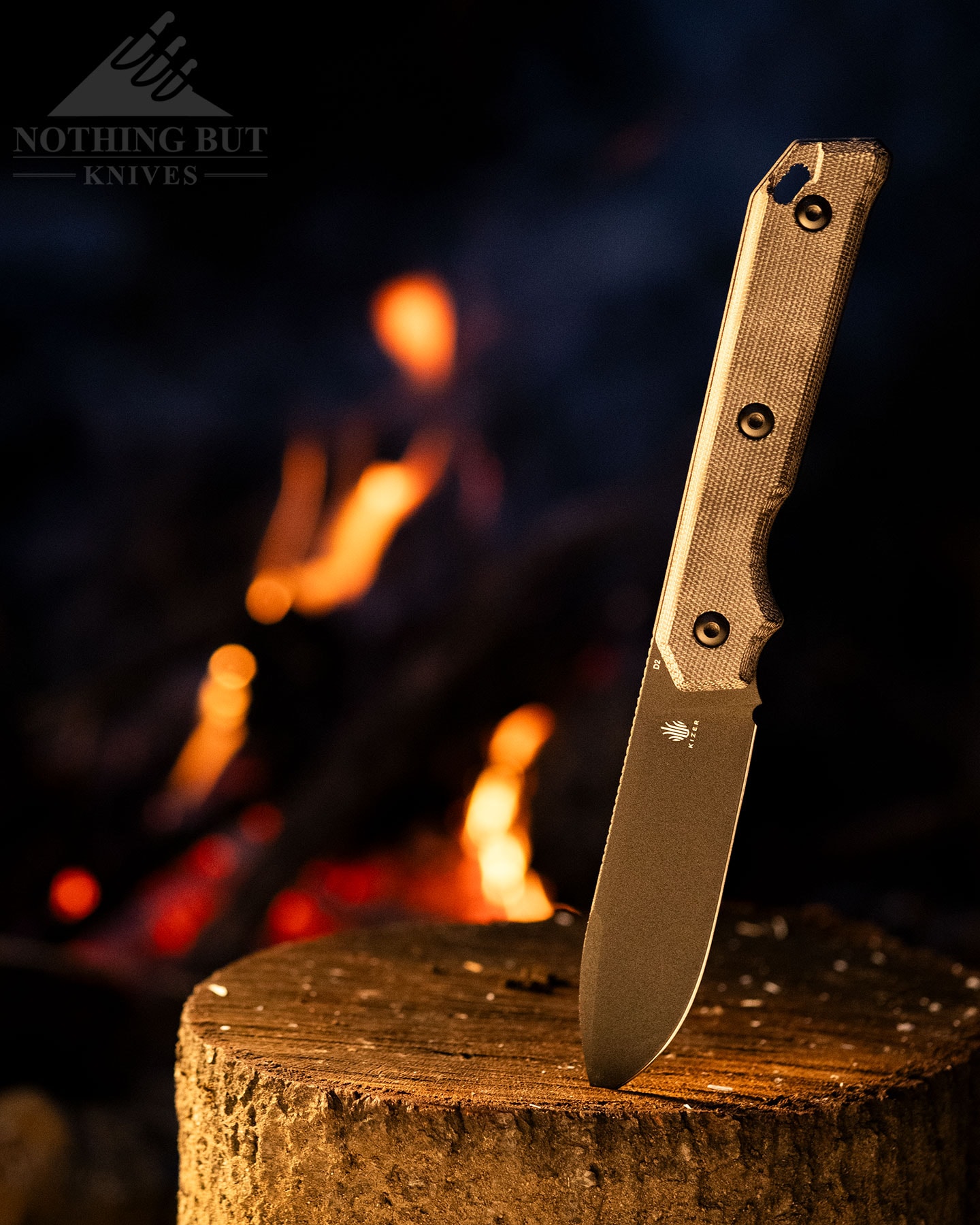 The Kizer Begleiter fixed blade knife shown here in front of a campfire with its blade tip sticking out of a piece of firewood. 