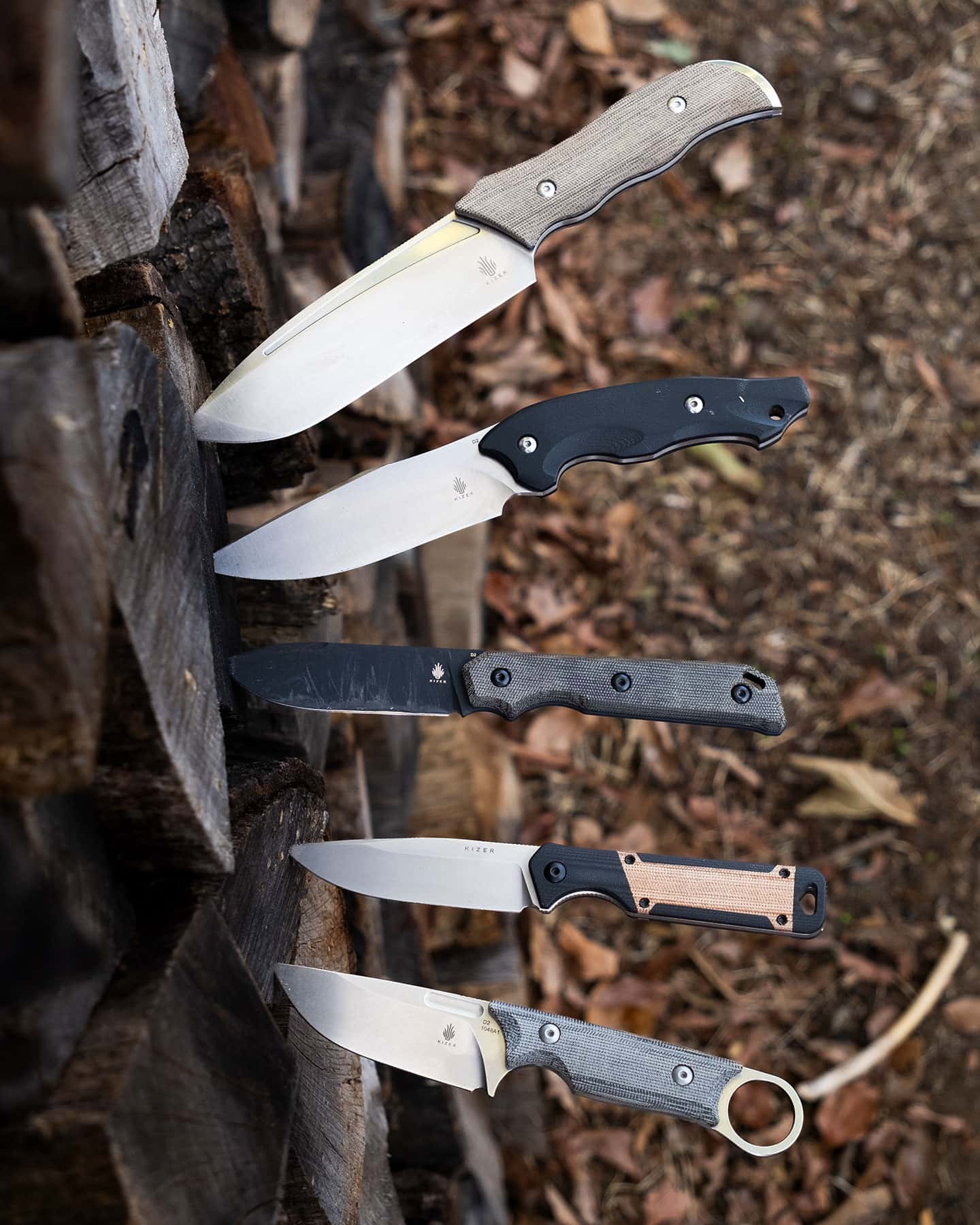 An overhead view of five Kizer fixed blade knives sticking out of firewood.