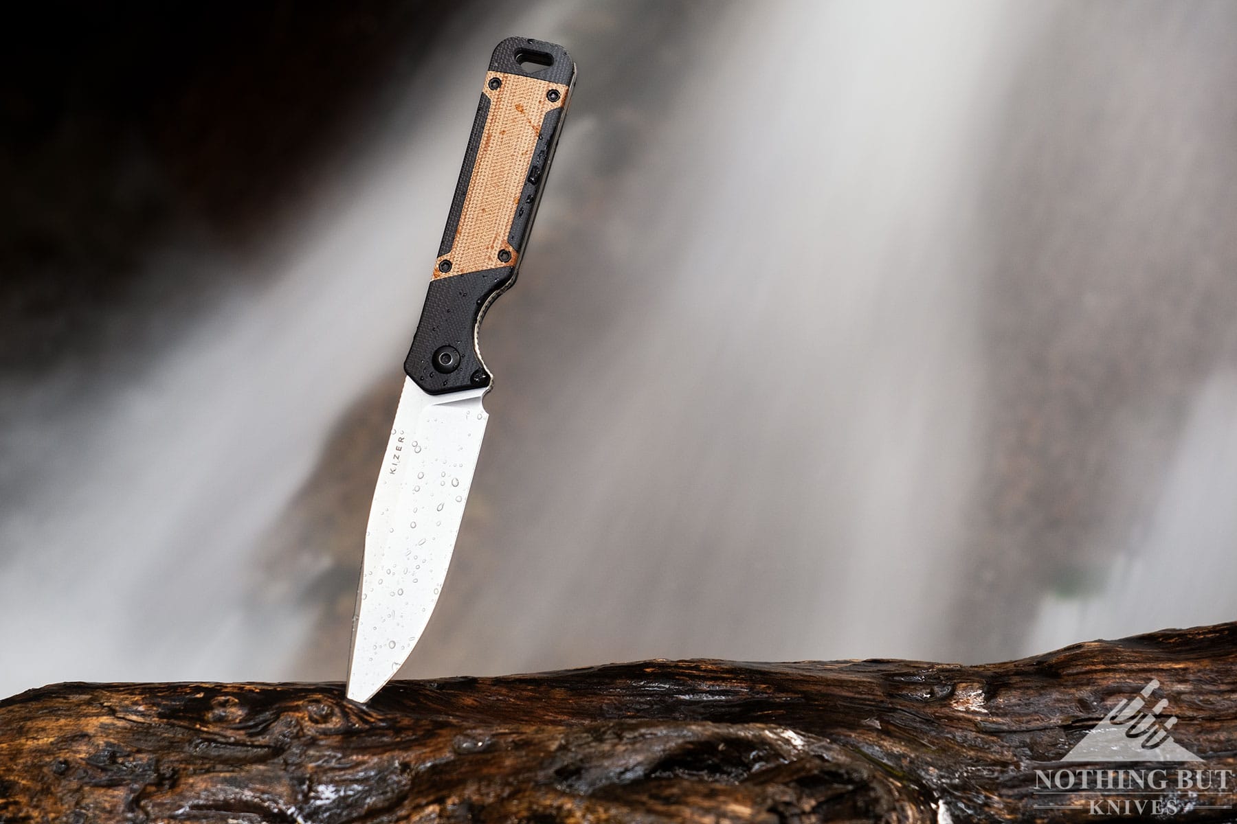 The Mizer Militaw Fixed Blade with its blade tip stuck in a piece of driftwood in front of a small waterfall. 