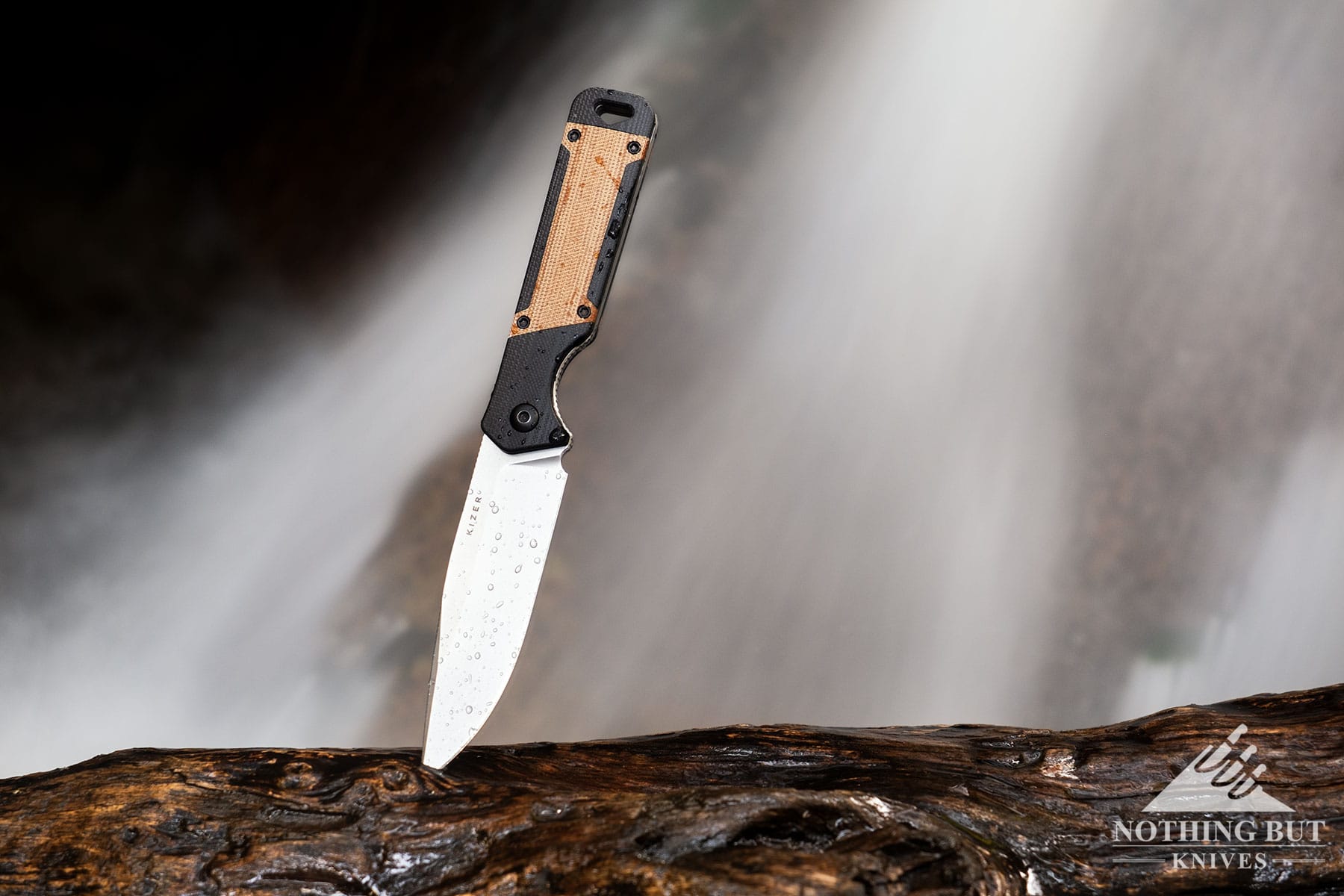 The Kizer Militaw fixed blade knife with its blade tip stuck in a piece of drift would next to a mountain stream. 