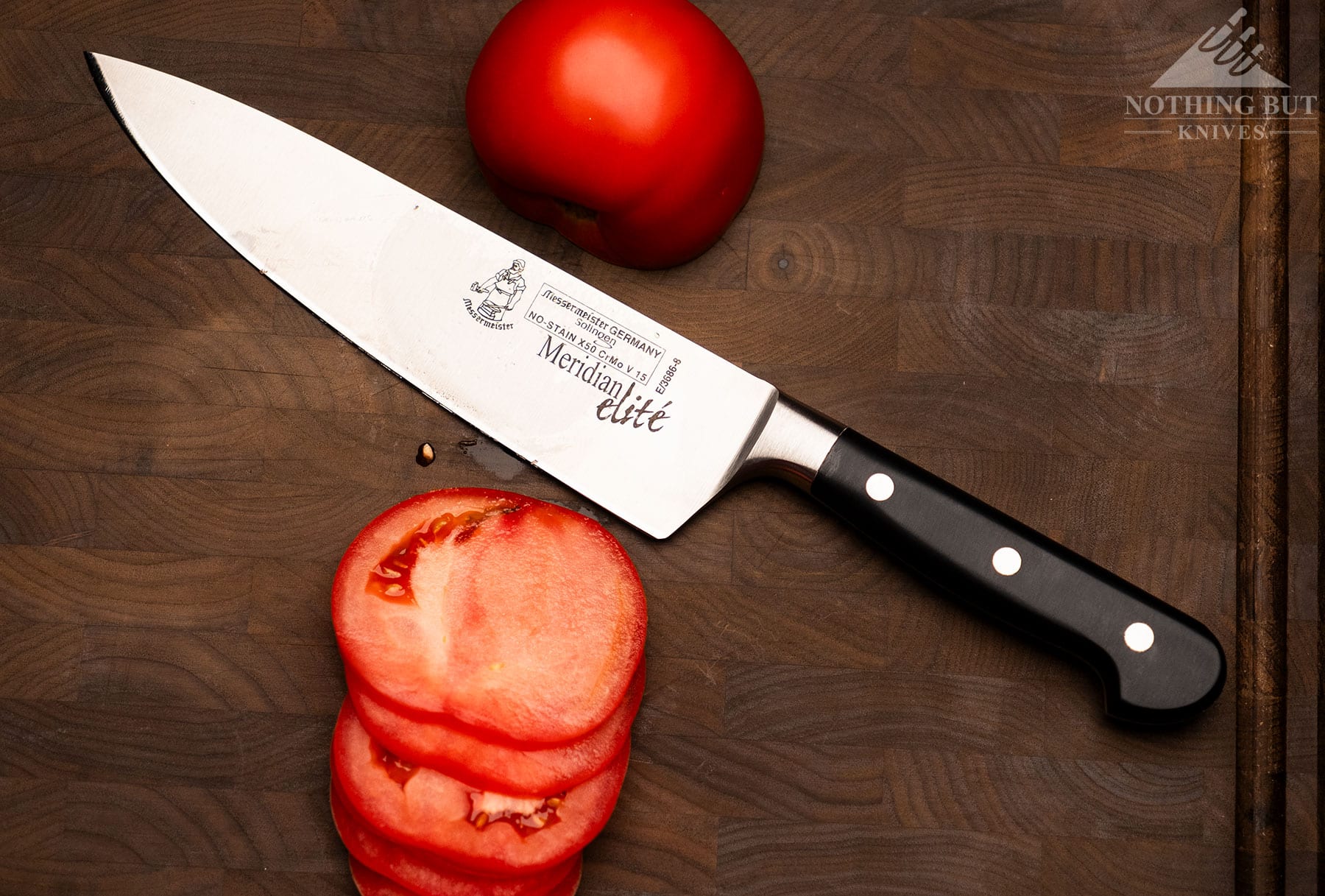 An overhead view of the Messermeister Meridian Elite 8-inch chef knife on a split grain cutting board between slices of a tomato it was just used to cut. 